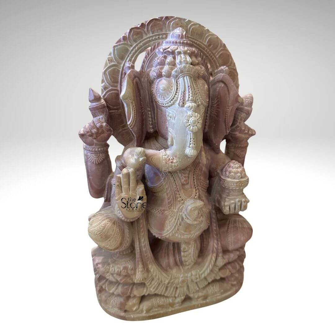 Pink Stone Ganpati Idol for Home 12" 6 A three-quarter front view photograph presents a premium twelve-inch Ganesha Murti meticulously hand-carved from a single block of natural pink stone. The statue depicts the revered elephant-headed Hindu deity Ganesha seated in a traditional meditative pose with one leg gently folded over the other resting upon a detailed multi-tiered lotus pedestal base. The pink stone exhibits a soft variegated hue ranging from pale rose to deeper mauve with subtle natural crystalline veining and inclusions throughout giving the idol a warm earthy and tactile texture. The sculpture is highly detailed showcasing the skill of the artisan in every contour. Ganesha is richly adorned with traditional deeply carved jewelry and ornaments all rendered with fine precision. He wears an elaborate multi-layered necklace armband anklets and a richly patterned crown that frames his serene face which features half-closed contemplative eyes and a gentle expression. His trunk curves gracefully to his left holding a single perfectly formed modak while his multiple hands bear symbolic attributes: his upper right hand holds a goad his upper left hand holds a noose his lower right hand is raised in the Abhaya mudra and his lower left hand holds a bowl overflowing with more modaks. The carving extends to his textured garments, which are draped and held by an ornate jeweled girdle and a decorative prabhavali with intricate floral and geometric motifs rises behind his head completing the divine iconography. This twelve-inch pink stone Ganesha statue is an exemplary piece of Indian stone carving art making it a perfect subject for an article on premium Ganesh Murtis. The compact size indicates its suitability for a home altar a prominent shelf or as a focal point in a smaller room bringing a sense of spiritual tranquility and cultural artistry to any interior space. The choice of pink stone adds a gentle inviting color to the decor while the detailed craftsmanship reflects a deep respect for traditional iconography. The photograph's clear focus on the statue's texture form and craftsmanship provides a comprehensive and engaging view for the viewer.