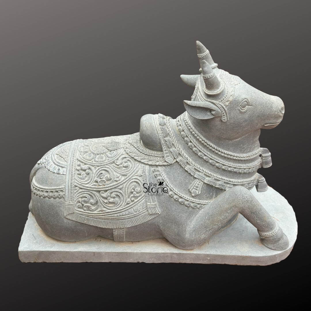 Nandikeshwara Stone Statue 45" 6 A detailed studio photograph showcases a meticulously hand-carved black stone Nandi bull statue positioned in a traditional recumbent pose with its right foreleg folded beneath its body. The statue exhibits a dark matte finish characteristic of premium blackstone and rests upon a plain rectangular stone base. Every surface of the Nandi is adorned with intricate carvings that highlight the skill of the artisan showcasing elaborate ornamentation. The Nandi’s head is turned slightly towards the viewer's right featuring finely detailed eyes ears and stylized horns. It is heavily decorated with multiple layers of detailed necklaces beads and bells including a prominent carved bell hanging from its neck all rendered in high relief directly from the black stone. The texture of the beads and metalwork is clearly visible. The body of the statue is covered by a beautifully ornate saddle blanket that drapes over its back and sides. The blanket features complex deep-relief floral motifs and geometric patterns bordered by detailed tassels and fringes. Below the blanket an intricately carved girth strap secures it ensuring the entire sculpture conveys a sense of rich ceremonial decoration. This premium blackstone statue embodies the traditional reverence for Nandi serving as a central focus for articles discussing spiritual art and temple sculpture.