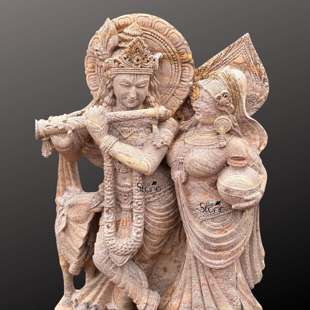 Radha Krishna Stone Statue with Cow 4ft 9 A close-up view of a magnificent 4-foot Radha Krishna Idol with Cow for Home intricately carved from warm reddish-brown sandstone reveals the delicate artistry against a dark gradient background. The statue captures Lord Krishna and Radha in an intimate and loving standing pose making it an ideal Idol for Home. Krishna distinguished by his elaborate crown and serene expression holds a flute to his lips seemingly in the act of playing with his head gently tilted towards Radha. Radha stands gracefully beside him adorned with a similar crown and traditional attire her gaze fixed on Krishna while she carries a pot of water or butter in her hand symbolizing her devotion and nurturing spirit. This detailed carving beautifully highlights their divine romance and interaction. The craftsmanship is exceptional, with every intricate detail defining their elaborate crowns delicate jewelry and flowing traditional garments. The sandstone's natural texture and subtle variations in color enhance the antique feel of the sculpture bringing out the tender expressions on their faces and the intricate patterns of their attire. Krishna's halo-like circular aureole with ornate patterns is partially visible behind him emphasizing his divine presence. A key element central to the article's title is the depiction of a small docile cow near Krishna's feet, symbolizing his role as a protector and caretaker of cattle and adding to the pastoral charm, which is highly desired for home idols. The figures stand on a multi-tiered intricately carved pedestal adorned with lotus petal designs and other traditional motifs providing a stable and dignified foundation. The depth and precision of the carving across the entire 4-foot statue are remarkable showcasing the artistry of the piece. This Sandstone Radha Krishna Idol with Cow is an exquisite example of religious artistry designed to evoke feelings of love devotion and peace perfectly aligning with an article focusing on such charming and symbolically rich Radha Krishna idols for home worship.
