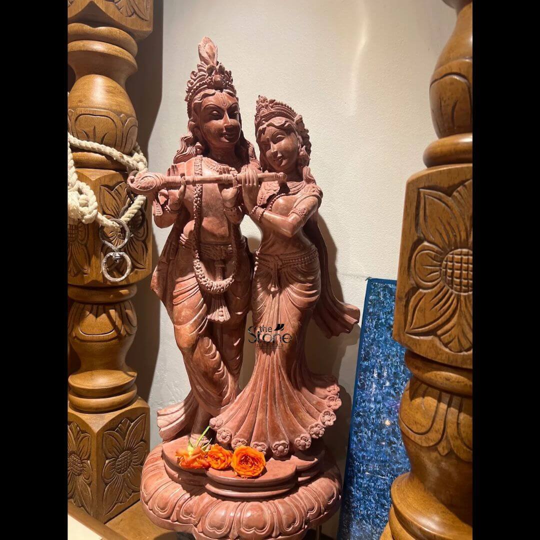 A medium shot captures a beautifully carved 20-inch Radha Krishna Pink Stone Statue positioned centrally on a wooden altar against a softly lit, neutral wall. The statue hewn from a single piece of fine-grained pink sandstone with warm, earthy hues, depicts the divine couple in a graceful tribhanga standing posture. Lord Krishna on the left is playing a bansuri while Radha standing intimately by his side with her hand on his shoulder gazes adoringly at him. Both figures are adorned with intricately sculpted traditional ornaments including layered necklaces armlets bracelets and elaborate crowns all rendered with high-relief detail that highlights the skill of the artisan. The statue rests on a round carved lotus-petal base adding to its spiritual significance. Several fresh vibrant orange rosebuds are placed as an offering on the base in front of the deities adding a touch of natural color and devotion to the composition. The stone itself has a smooth polished texture that catches the warm light emphasizing the soft contours of their faces and the flow of their garments. The deep pink color of the stone gives the sculpture a warm inviting glow making it a captivating focal point. This exquisite pink stone statue of Radha and Krishna is a perfect representation of divine love and is a sought-after piece of art for spiritual and decorative purposes particularly among the Indian diaspora in Kuala Lumpur, Malaysia. The use of sandstone known for its durability and aesthetic appeal, makes it a long-lasting addition to a home shrine or living space. The craftsmanship and the serene expressions of the deities convey a sense of peace and devotion embodying the rich cultural heritage of India. The size and detailed carving make it an ideal centerpiece for worship or as a cultural artifact in a Malaysian home.