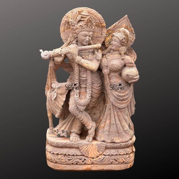 A magnificent 4-foot Sandstone Radha Krishna statue prominently featuring a cow stands gracefully against a dark gradient background radiating divine love and harmony. The statue carved from warm reddish-brown sandstone depicts Lord Krishna and Radha standing lovingly side-by-side. Krishna distinguished by his elaborate crown and serene expression holds a flute to his lips poised to play a melodious tune. Radha stands gracefully beside him adorned with a crown and traditional attire carrying a pot of water or butter symbolizing her devotion and nurturing spirit. This detailed carving beautifully captures their divine romance and pastoral setting. The craftsmanship is exquisite with intricate details defining their elaborate crowns delicate jewelry and flowing traditional garments. The sandstone's natural texture and subtle variations in color enhance the antique feel of the sculpture bringing out the tender expressions on their faces and the intricate patterns of their attire. A halo-like circular aureole with ornate patterns frames Krishna's head further emphasizing his divine presence. A key element central to the article's title is the inclusion of a small docile cow depicted near Krishna's feet symbolizing his role as a protector and caretaker of cattle and adding to the pastoral charm. The entire ensemble rests on a multi-tiered intricately carved pedestal adorned with lotus petal designs and other traditional motifs providing a stable and dignified foundation. The depth and detail of the carving across the entire 4-foot statue are remarkable showcasing the artistry of the piece. This Sandstone Radha Krishna statue with its endearing depiction of a cow is an exquisite example of religious artistry designed to bring blessings love and aesthetic beauty to any home or temple making it an ideal focal point for devotion perfectly aligning with an article focusing on such charming Radha Krishna statues with cows.
