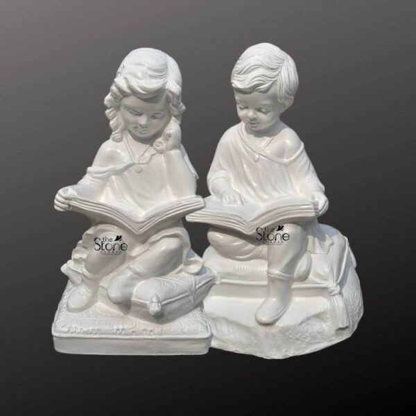 A three-quarter view photograph captures a pair of beautifully detailed matte white garden statues depicting a young boy and girl seated and engrossed in reading. The statues are made of durable fiberglass giving them a smooth stone-like appearance while retaining the lightweight benefits of a modern composite material. They are positioned side-by-side against a softly graduated dark grey background which highlights their bright clean color. Both figures are seated cross-legged on individual textured cushions adding a cozy natural element to their poses. The girl on the left is seated on a cushion with a subtle decorative border her head tilted slightly in concentration as she holds an open book on her lap. Her curly hair is sculpted with care and she wears a simple dress. The boy on the right is also seated on a separate cushion looking down at his own open book with focused attention. He wears a tunic-style top and trousers, with his short hair neatly styled. The books held by both children are rendered with visible pages and covers adding realism to the scene. This delightful Reading Boy & Girl Fibre Garden Statue Set is specifically designed as charming decor for a garden or outdoor reading nook. The use of fiberglass construction ensures these statues are weather-resistant and portable making them ideal for placement among garden foliage on a patio or near a bench. The theme of the statues evokes a sense of childhood innocence and a love for learning making them a heartwarming addition to any outdoor space. Their detailed sculpting and bright white finish create a striking contrast against green plants and colorful flowers.