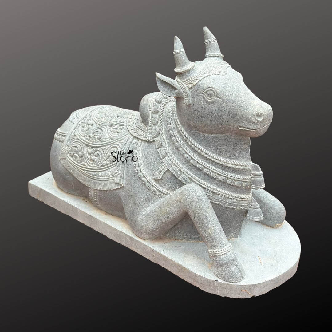 Nandikeshwara Stone Statue 45" 7 A detailed studio photograph captures a meticulously hand-carved blackstone Nandi bull idol presented in a traditional recumbent pose. The sacred statue resting on a plain rectangular stone base, exhibits the dark matte grey finish characteristic of premium blackstone. Nandi is depicted lying down with its right foreleg gracefully folded beneath its body facing towards the viewer's right with a serene attentive expression. The entire body of the Nandi idol is richly adorned with elaborate deeply carved ornamentation. Its face features detailed eyes ears and stylized horns. Multiple layers of intricately carved necklaces composed of varied bead sizes and bell pendants drape around its neck with the texture of the stone mimicking heavy jewelry. A prominent ornate carved bell hangs centrally from these layers. The Nandi’s back is covered by a beautifully patterned saddle blanket secured by an intricately carved girth strap. The blanket itself is covered in dense detailed floral motifs and geometric borders with tassels and fringe carved along the lower edge. Every surface of the sculpture showcases the high level of craftsmanship emphasizing its role as a sacred temple idol and a revered object of spiritual art. The statue is set against a smooth dark grey gradient background isolating the figure for clear visibility.