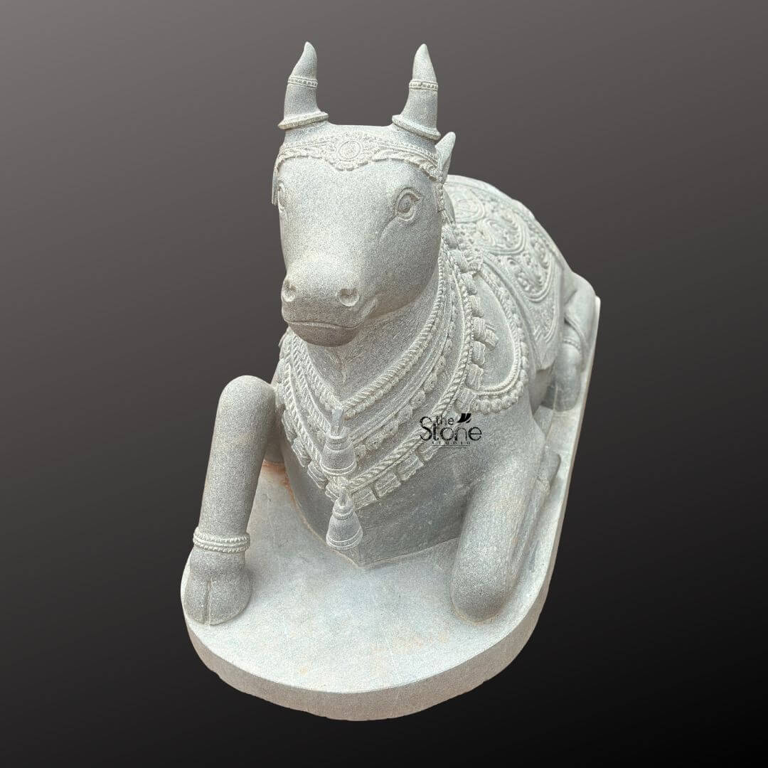 Nandikeshwara Stone Statue 45" 8 A detailed elevated three-quarter view studio photograph showcases a magnificent hand-carved blackstone Nandi bull statue. The sacred idol is presented in a traditional recumbent pose resting on a plain elevated rectangular stone plinth against a smooth dark grey gradient background that isolates the figure. The statue exhibits the dark rich and matte finish characteristic of premium blackstone with its surface meticulously worked to create both smooth forms and deep-relief textures. Nandi is depicted with its right foreleg gracefully folded beneath its body and its head turned slightly to the viewer's left gazing forward with a serene and watchful expression. The Nandi statue is richly adorned with elaborate ornamentation carved directly from the stone. Its head features detailed eyes prominent ears and intricately carved horns with a delicate headpiece resting on its forehead. Multiple layers of finely carved beaded necklaces, chains, and bell pendants drape around its neck, simulating heavy temple jewelry with the texture of the beads and bells clearly defined in high relief. The sculpture conveys a sense of deep spiritual reverence through its detailed craftsmanship. The body of the Nandi is covered by an intricately patterned saddle blanket that drapes over its back and sides. The blanket is adorned with dense floral motifs geometric patterns and a textured border finished with carved tassels and fringes along the lower edge. Below the blanket a detailed girth strap secures it ensuring the sculpture embodies the ceremonial decoration befitting a sacred temple statue. The entire piece exemplifies the high quality of traditional Indian stone carving making it a fitting subject for articles discussing sacred art and temple iconography.