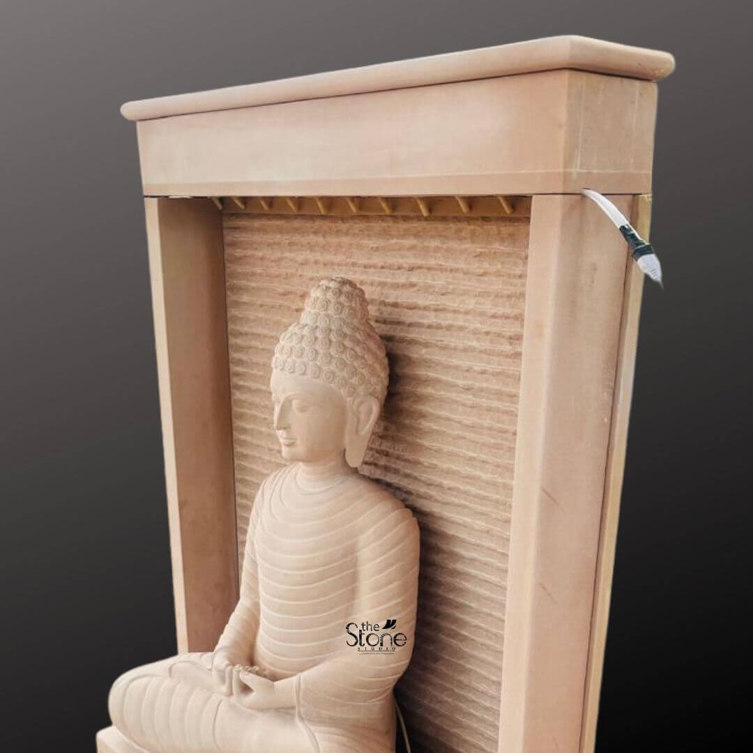 Outdoor Buddha Stone Water Fountain 4.5ft 7 A four-and-a-half-foot tall Buddha statue is the tranquil centerpiece of a contemporary off-white sandstone water fountain presented here in a clean studio setting against a dark grey background. The statue hand-carved from the beautiful light beige-to-grey sandstone depicts the Buddha in a seated meditative pose on a raised pedestal with hands resting in the lap in the dhyana mudra of deep contemplation. The profile view emphasizes the serene facial expression with downcast eyes a gentle smile and tightly curled hair leading to a ushnisha. The flowing folds of the monastic robes are subtly rendered showcasing the natural textured porous surface of the sandstone material. The Buddha statue is enclosed within a monolithic frame-like sandstone structure creating a modern wall-fountain design. Water is subtly distributed to flow like a gentle curtain down the textured back panel directly behind the Buddha pooling into the large monolithic rectangular basin at the base. The natural earthy hue of the sandstone contrasts beautifully with the dark background making the statue pop and highlighting its intricate details and substantial presence. The overall design is a harmonious blend of sacred art and the calming element of cascading water. This four-and-a-half-foot Buddha Fountain Off White Sandstone Statue is an ideal illustration for an article discussing Sandstone Buddha Fountain demonstrating how such a piece can transform an indoor or outdoor space into a peaceful sanctuary. Its large scale ensures it becomes a commanding focal point while the durable sandstone construction makes it suitable for enduring elements as a long-lasting garden ornament. The combination of traditional Buddhist iconography with a modern water feature design embodies a sense of calm contemplation and aesthetic beauty perfect for creating a dedicated space for meditation and reflection. The statue's presence evokes a sense of sacred history and spiritual peace enhancing the environment with its serene and contemplative form.