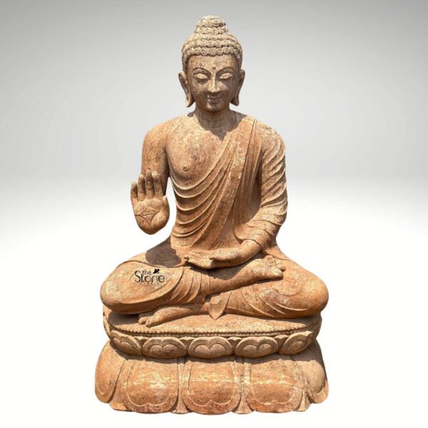 A magnificent 3-foot Sandstone Buddha Statue in Abhaya Mudra stands prominently against a clean light grey background radiating peace and reassurance. The statue carved from a warm reddish-brown sandstone depicts Buddha in a serene meditative posture seated in the full lotus position symbolizing deep concentration and spiritual enlightenment. His right hand is raised to shoulder height with the palm facing outwards fingers extended upwards forming the Abhaya Mudra the gesture of fearlessness blessing and protection which is the central theme for the article. His left hand rests gently in his lap palm upwards signifying contemplation and receptivity. The natural earthy hue of the sandstone gives the sculpture an authentic ancient feel highlighting its intricate details. The Buddha's facial features are exquisitely carved radiating profound peace and wisdom. His eyes are gently closed or cast downwards in contemplation and a subtle serene smile graces his lips. His head is adorned with tightly coiled hair culminating in the ushnisha the cranial bump symbolizing his supreme spiritual knowledge and enlightenment. The flowing drapery of his robes is sculpted with soft elegant folds emphasizing the texture of the sandstone and contributing to the statue's graceful and tranquil form. The weathered appearance of the sandstone adds to its charm and gives it a timeless quality. The Buddha is seated upon a beautifully sculpted double-lotus pedestal with each petal intricately carved symbolizing purity spiritual growth and the emergence of enlightenment from the mundane. The robust and detailed base supports the statue with dignity making it a focal point. This 3-foot Sandstone Buddha statue with its clear depiction of the Abhaya Mudra is an exquisite example of religious artistry designed to bring blessings peace and reassurance to any indoor or outdoor space perfectly aligning with an article focusing on Buddha statues specifically in the Abhaya Mudra.