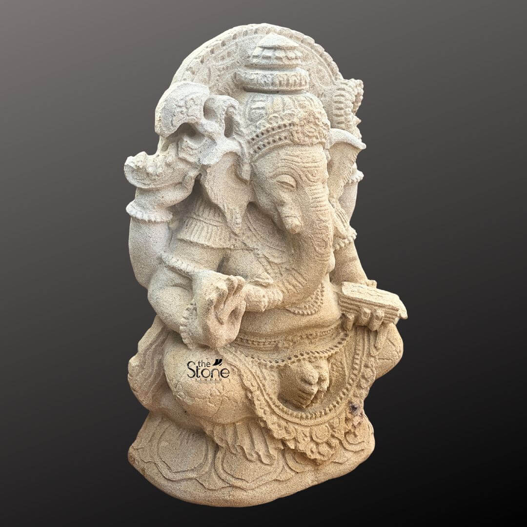 Indonesian Ganesha Idol for Home 2ft 5 A detailed three-quarter view photograph captures a hand-carved sandstone Ganesha statue approximately two feet tall. The sculpture depicts the elephant-headed Hindu deity seated in a traditional Lalitasana pose with his left leg folded and his right leg pendent resting on a multi-tiered lotus-carved pedestal. The statue is hewn from a light-beige sandstone that exhibits a textured granular surface enhancing its antique look and weathered patina. Ganesha is adorned with intricate deeply carved ornaments typical of classical Indian sculpture, including elaborate necklaces, armlets, anklets, and a richly detailed crown featuring floral and geometric motifs. A decorative halo with a flaming border frames his head and torso, adding to the divine presence. The four-armed deity holds symbolic attributes in his hands all rendered with fine detail. His upper right hand holds an axe while his upper left hand holds a broken tusk. His lower right hand is raised in the Abhaya mudra, a gesture of blessing and fearlessness, and his lower left hand holds a modak towards which his trunk gently curves. The sculptor has paid meticulous attention to the texture of his dhoti the folds of which are clearly defined beneath a jeweled girdle. The figure's posture is serene and balanced with a gentle expression on his face and eyes half-closed in meditation characteristic of traditional devotional art. This antique-look sandstone Ganesha statue is an exceptional example of stone carving craftsmanship making it highly relevant as a visual reference for an article on the subject. The weathered finish and the specific iconographic details—such as the number of arms the specific attributes held, and the ornamental style—provide valuable insights into the cultural and artistic traditions of Ganesha statues. The statue's size and detailed carvings make it a suitable piece for home shrines, garden focal points or as a collector's item. The photograph highlights the porous texture and natural color variations of the sandstone, showcasing the material's inherent beauty and its suitability for intricate sculptural work.