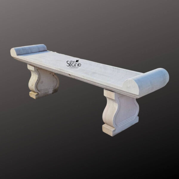 A six-foot-long sandstone garden bench stands against a seamless dark grey studio background showcasing its elegant classical design. The bench is hand-carved from natural light beige sandstone exhibiting a beautifully textured porous surface that adds an authentic rustic charm. Its long flat seat is supported by two substantial decoratively sculpted legs. These legs feature a graceful S-curve or hourglass profile resting on sturdy block-like bases that anchor the entire piece. The craftsmanship is evident in the smooth yet hand-hewn finish of the stone which catches the light and highlights its natural variations in color and texture. The design of this sandstone bench is distinctly traditional inspired by historical European garden furniture. The seat itself is not plain it terminates at both the left and right ends with large perfectly formed scrolled volutes or rolled ends. These sculptural elements not only provide comfortable armrests but also define the bench's aesthetic lending it a sophisticated and timeless appeal. The overall form is balanced and substantial suggesting durability and a permanent place within a garden landscape. The stone appears heavy and solid capable of withstanding the elements for decades. This six-foot sandstone bench is the perfect embodiment of a functional and decorative Sandstone Garden Bench. Its generous length provides ample seating for multiple people while its robust stone construction makes it an ideal choice for outdoor settings like patios courtyards or nestled within garden pathways. The light neutral color of the sandstone harmonizes beautifully with surrounding greenery flowers and other natural elements. This bench is not just a piece of furniture it is a sculptural addition to a garden designed to offer a comfortable and contemplative spot to rest and enjoy the beauty of the outdoors.