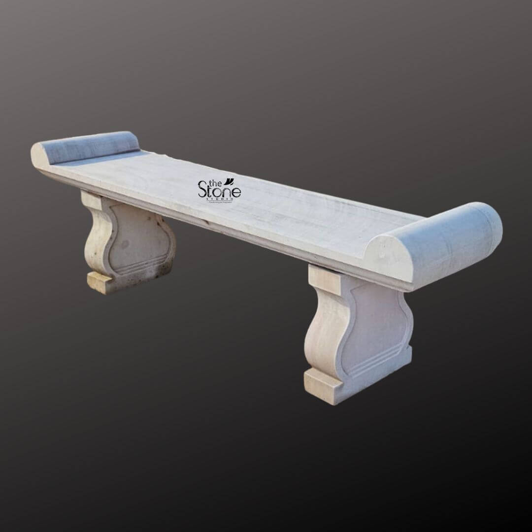 Sandstone Garden Bench 6ft 1 A six-foot-long sandstone garden bench stands against a seamless dark grey studio background showcasing its elegant classical design. The bench is hand-carved from natural light beige sandstone exhibiting a beautifully textured porous surface that adds an authentic rustic charm. Its long flat seat is supported by two substantial decoratively sculpted legs. These legs feature a graceful S-curve or hourglass profile resting on sturdy block-like bases that anchor the entire piece. The craftsmanship is evident in the smooth yet hand-hewn finish of the stone which catches the light and highlights its natural variations in color and texture. The design of this sandstone bench is distinctly traditional inspired by historical European garden furniture. The seat itself is not plain it terminates at both the left and right ends with large perfectly formed scrolled volutes or rolled ends. These sculptural elements not only provide comfortable armrests but also define the bench's aesthetic lending it a sophisticated and timeless appeal. The overall form is balanced and substantial suggesting durability and a permanent place within a garden landscape. The stone appears heavy and solid capable of withstanding the elements for decades. This six-foot sandstone bench is the perfect embodiment of a functional and decorative Sandstone Garden Bench. Its generous length provides ample seating for multiple people while its robust stone construction makes it an ideal choice for outdoor settings like patios courtyards or nestled within garden pathways. The light neutral color of the sandstone harmonizes beautifully with surrounding greenery flowers and other natural elements. This bench is not just a piece of furniture it is a sculptural addition to a garden designed to offer a comfortable and contemplative spot to rest and enjoy the beauty of the outdoors.