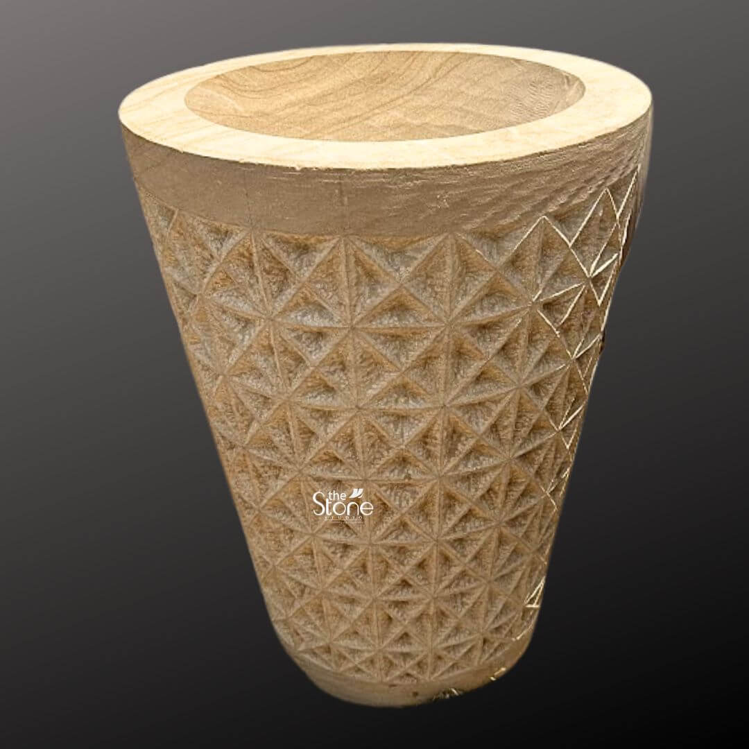 Natural Sandstone Planter Pot 2ft 6 A full-view photograph of a magnificent 2-foot tall sandstone landscape planter pot presented against a dark neutral gray gradient background that accentuates its form. The planter is hand-carved from a solid block of warm textured beige sandstone showcasing a rich natural grain and tonal variations that highlight its geological origin. It is fashioned into a classic slightly tapered cylindrical shape providing a robust and stable structure for large plants. The overall impression is one of rustic elegance and durable craftsmanship perfectly suited for enhancing any garden or outdoor area. The central body of the sandstone landscape planter pot is adorned with an intricate deeply incised geometric pattern. This pattern consists of a repeating tessellation of diamond and triangular facets creating a textured three-dimensional surface that catches the light and adds significant visual depth. The precision and detail of the carving demonstrate a high level of artisanal skill turning the functional object into a piece of sculptural art. The natural porosity of the sandstone is clearly visible emphasizing its suitability for outdoor environments where it can withstand the elements while allowing soil and plants to breathe. This substantial 2-foot stone planter pot is a versatile and attractive choice for any landscaping project. Its scale and intricate design make it an ideal focal point for a patio deck or garden path providing a sturdy and stylish home for large flowering plants or small shrubs. The image clearly conveys the texture scale and elaborate geometric carving of the planter making it a perfect visual reference for an article discussing natural stone elements in garden design and the beauty of hand-carved sandstone products.