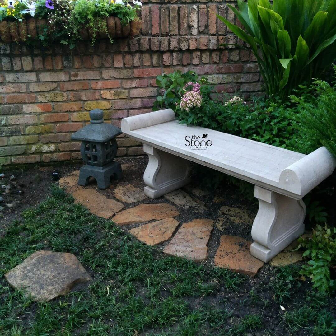 Sandstone Garden Bench 6ft 3 A six-foot-long sandstone outdoor bench is positioned as a tranquil focal point within a secluded garden nook. The bench is hand-carved from natural light beige sandstone showcasing a beautifully textured porous surface that adds an authentic rustic charm. Its long flat seat is supported by two substantial decoratively sculpted legs. These legs feature a graceful S-curve or hourglass profile resting on sturdy block-like bases that anchor the entire piece. The craftsmanship is evident in the smooth yet hand-hewn finish of the stone which catches the natural daylight and highlights its inherent variations in color and texture. The design of this sandstone bench is distinctly classical inspired by historical European garden furniture. The seat itself is not plain it terminates at both the left and right ends with large perfectly formed scrolled volutes or rolled ends. These sculptural elements not only provide comfortable armrests but also define the bench's aesthetic lending it a sophisticated and timeless appeal. The overall form is balanced and substantial suggesting durability and a permanent place within a garden landscape. The stone appears heavy and solid capable of withstanding the elements for decades. This six-foot sandstone bench is the perfect embodiment of a functional and decorative Sandstone Outdoor Bench. Its generous length provides ample seating for multiple people while its robust stone construction makes it an ideal choice for outdoor settings like patios courtyards or nestled within garden pathways. The light neutral color of the sandstone harmonizes beautifully with surrounding greenery flowers and other natural elements. The setting here which includes a weathered brick wall lush plants a stone path and a decorative Japanese-style lantern perfectly illustrates how this bench can be integrated into a variety of garden styles to create a peaceful and inviting sanctuary.
