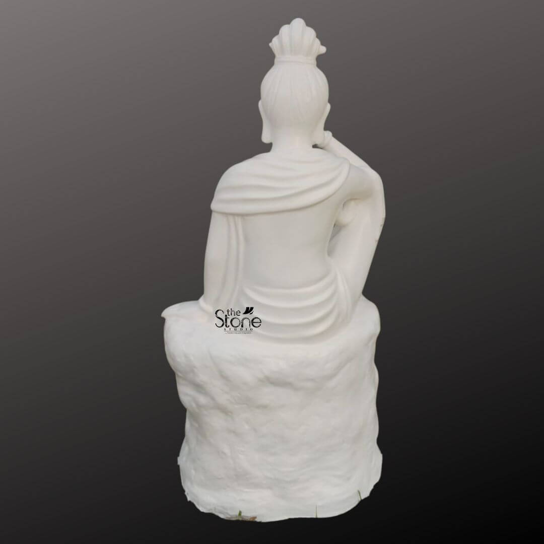Seated Kuan Yin Statue 3ft 5 A rear view of a 3 feet fibre Seated Avalokiteshvara statue is shown against a smooth dark grey gradient background presenting the sculpture in a clean product-style format. The statue depicts Kuan Yin also known as Avalokiteshvara or Guanyin seated gracefully on a textured rock-like pedestal. Seen from behind the figure has a calm poised posture with the upper body upright and one arm raised toward the face suggesting a quiet meditative seated pose. The back profile reveals the balanced proportions of the sculpture and helps viewers understand the full form of the statue beyond the front-facing devotional expression. The white fibre Kuan Yin statue features softly sculpted drapery flowing across the shoulders and down the back with elegant robe folds wrapping around the body and settling naturally over the seated form. The hairstyle is gathered into a high bun or crowned topknot adding to the statue’s refined spiritual appearance while the elongated ears and gently modelled shoulders enhance the graceful silhouette. The textured pedestal beneath the seated figure resembles carved stone giving the sculpture visual depth and a grounded natural base. This back-angle view highlights the craftsmanship smooth finish and thoughtful detailing of the Seated Avalokiteshvara statue from all sides. This image connects well to an article about a Seated Avalokiteshvara Statue by showing the back view of a 3 feet fibre Kuan Yin statue helping readers appreciate its sculptural design material and decorative versatility. It presents the statue as a suitable choice for home décor meditation spaces spiritual corners temples wellness interiors or peaceful garden-inspired settings where a full 360-degree aesthetic matters. The serene posture flowing robe details and clean white finish reflect the compassionate and meditative essence of Avalokiteshvara making this fibre statue a meaningful and elegant addition to devotional and decorative spaces.