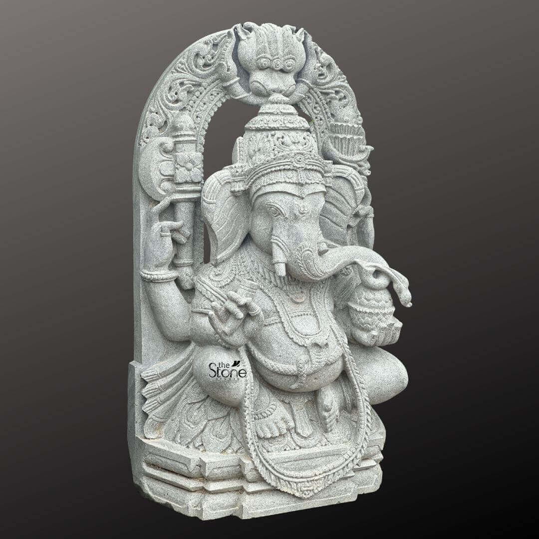 Ganesha Statue with Makara Torana 3ft 6 A magnificent 3-foot Seated Ganesha Statue with Makara Torana intricately carved from what appears to be a light grey stone stands powerfully against a dark gradient background. The statue depicts Lord Ganesha in a regal seated posture exuding benevolence wisdom and an aura of divine grace. His iconic elephant head is finely sculpted complete with large ears expressive eyes and his distinctive broken tusk delicately poised to write or interact. He is adorned with an elaborate crown multiple layers of traditional jewelry including necklaces armlets and a sacred thread draped across his chest all rendered with exceptional detail in the light grey stone. Ganesha is portrayed with four arms holding his customary attributes: a modak in one hand an axe or goad in another and his lower right hand extended in a gesture of blessing offering reassurance and protection. The intricate details on his garments the patterns on his halo and the careful sculpting of his prominent pot belly are all clearly visible showcasing the high artistry of the carving. The light grey stone emphasizes these textures and details lending a serene and timeless elegance to the deity and suggesting a weathered ancient feel that speaks to its profound presence. The statue is dramatically framed by a grand ornate archway clearly a Makara Torana which features exquisite mythical sea-creature motifs at its base and intricate traditional carvings along its entire span and supporting pillars. The top of the arch often features a Kirtimukha (a fierce lion-like face a common protective motif. This entire elaborate structure rests on a multi-tiered base also carved from the same light grey stone providing stability and enhancing its grand temple-like presence. This 3-foot Seated Ganesha Statue with its impressive Makara Torana is an exquisite example of religious artistry designed to bring blessings peace and aesthetic grandeur to any space perfectly aligning with an article focusing on such a revered Ganesha sculpture.