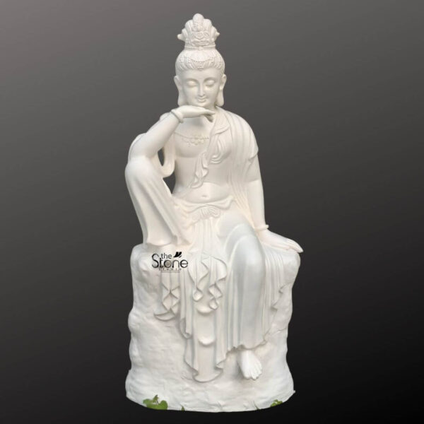 A 3 feet fibre Seated Kuan Yin Statue is shown in a clean product-style presentation against a smooth dark grey background making the sculpture the clear focal point of the image. The statue depicts Kuan Yin the bodhisattva of compassion seated gracefully on a textured rock-like base in a relaxed and contemplative pose. One leg is raised with the arm resting gently across the knee while the other leg falls softly downward creating an elegant balanced silhouette. The face has a calm serene expression with closed eyes and delicate features conveying peace kindness and spiritual stillness. The white fibre Kuan Yin statue is richly detailed with flowing robes draped in soft folds across the body and cascading down the pedestal-like base. Fine ornamental elements can be seen in the crown or headdress along with subtle jewellery details around the neck and wrists enhancing the artistic and devotional character of the sculpture. The smooth white finish gives the statue a refined graceful appearance while the sculpted rock seat adds depth and visual contrast. The combination of the compassionate expression elegant posture and detailed craftsmanship makes the statue visually striking yet soothing. This image connects well to an article about a Seated Kuan Yin Statue by clearly highlighting the form material and decorative appeal of a 3 feet fibre Kuan Yin statue. It presents the sculpture as a beautiful choice for spiritual décor meditation spaces peaceful interiors garden corners temple settings or serene entrance styling. The overall look emphasizes the statue’s devotional beauty calm presence and decorative versatility making it an ideal representation of a seated Kuan Yin fibre statue for anyone seeking a symbol of compassion peace and elegant spiritual art.
