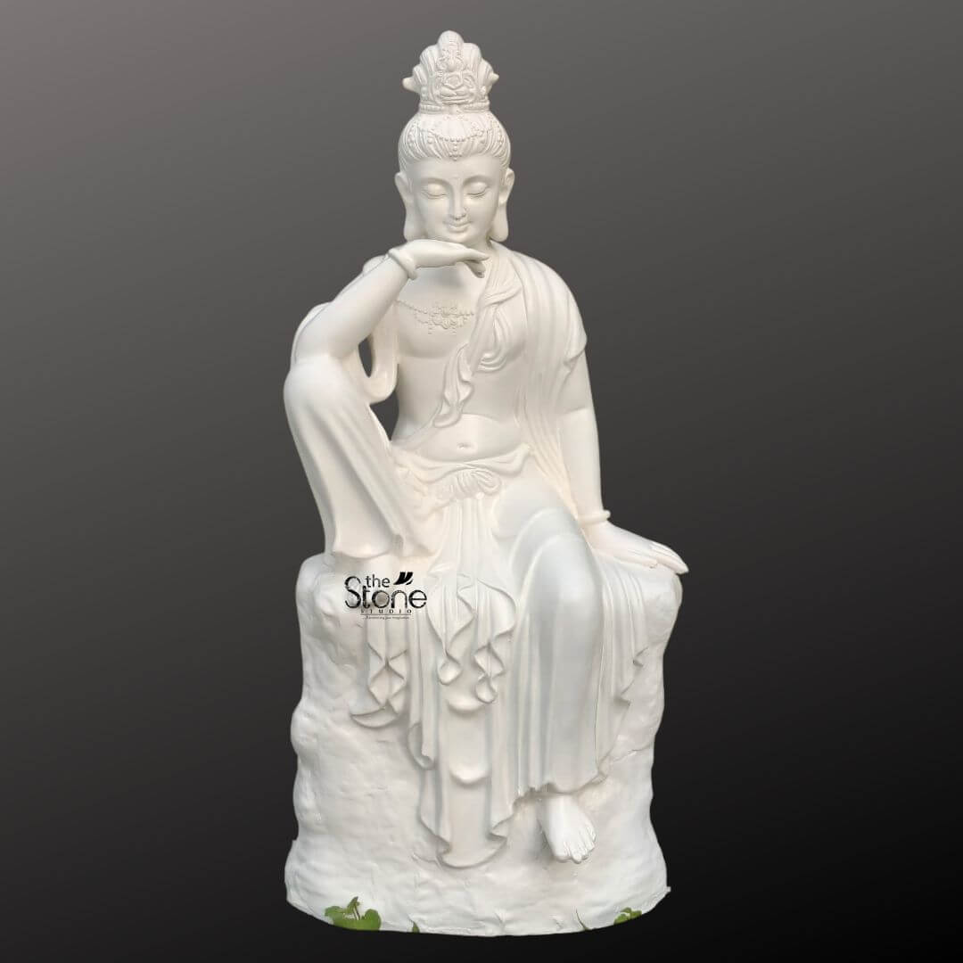 Seated Kuan Yin Statue 3ft 1 A 3 feet fibre Seated Kuan Yin Statue is shown in a clean product-style presentation against a smooth dark grey background making the sculpture the clear focal point of the image. The statue depicts Kuan Yin the bodhisattva of compassion seated gracefully on a textured rock-like base in a relaxed and contemplative pose. One leg is raised with the arm resting gently across the knee while the other leg falls softly downward creating an elegant balanced silhouette. The face has a calm serene expression with closed eyes and delicate features conveying peace kindness and spiritual stillness. The white fibre Kuan Yin statue is richly detailed with flowing robes draped in soft folds across the body and cascading down the pedestal-like base. Fine ornamental elements can be seen in the crown or headdress along with subtle jewellery details around the neck and wrists enhancing the artistic and devotional character of the sculpture. The smooth white finish gives the statue a refined graceful appearance while the sculpted rock seat adds depth and visual contrast. The combination of the compassionate expression elegant posture and detailed craftsmanship makes the statue visually striking yet soothing. This image connects well to an article about a Seated Kuan Yin Statue by clearly highlighting the form material and decorative appeal of a 3 feet fibre Kuan Yin statue. It presents the sculpture as a beautiful choice for spiritual décor meditation spaces peaceful interiors garden corners temple settings or serene entrance styling. The overall look emphasizes the statue’s devotional beauty calm presence and decorative versatility making it an ideal representation of a seated Kuan Yin fibre statue for anyone seeking a symbol of compassion peace and elegant spiritual art.