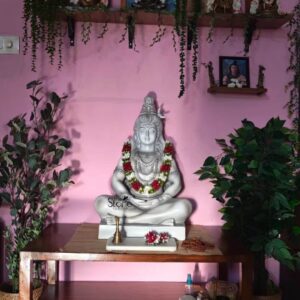 A reverent eye-level photograph captures a beautifully decorated home shrine centered around a 2 Feet Shiva Fibre Statue positioned against a warm pink-painted wall in a Puducherry home. The statue depicts Lord Shiva in a serene meditative padmasana pose hand-painted in a matte white finish with subtle grey shading to define the musculature and traditional ornamentation. He is adorned with a vibrant fresh garland of red and white flowers which drapes over his chest and shoulders contrasting with the white material. A miniature brass cobra hood is integrated above his head and the sacred river Ganges flows from his matted hair showcasing intricate detailing. The idol rests on a simple elegant wooden altar table creating a peaceful focal point for worship. This Fibre Shiva Idol is a substantial piece measuring approximately 2 feet in height making it perfectly scaled for a home puja room or personal altar space without being overpowering. The use of fiberglass construction ensures the statue is lightweight yet durable allowing for easier handling and placement compared to a traditional stone sculpture of similar size while still retaining a high level of detailed craftsmanship in the facial features and jewelry. The idol is flanked by two large realistic artificial potted plants housed in woven baskets adding a touch of nature and symmetry to the sacred space. Above the altar a wall-mounted wooden shelf displays various framed Hindu deities and spiritual images, including depictions of Lakshmi Vishnu and a larger colorful painting of Shiva and Parvati on Nandi all adorned with strings of artificial green vines. The lower shelf of the altar table holds essential puja items including a small brass oil lamp prayer beads and various ritual offerings emphasizing the idol's functional role in daily worship. The entire composition highlights the significance of this 2-foot fibre Shiva idol as an accessible and spiritually meaningful addition to a home blending traditional devotion with modern materials and reflecting a common style of home shrine found in Puducherry.