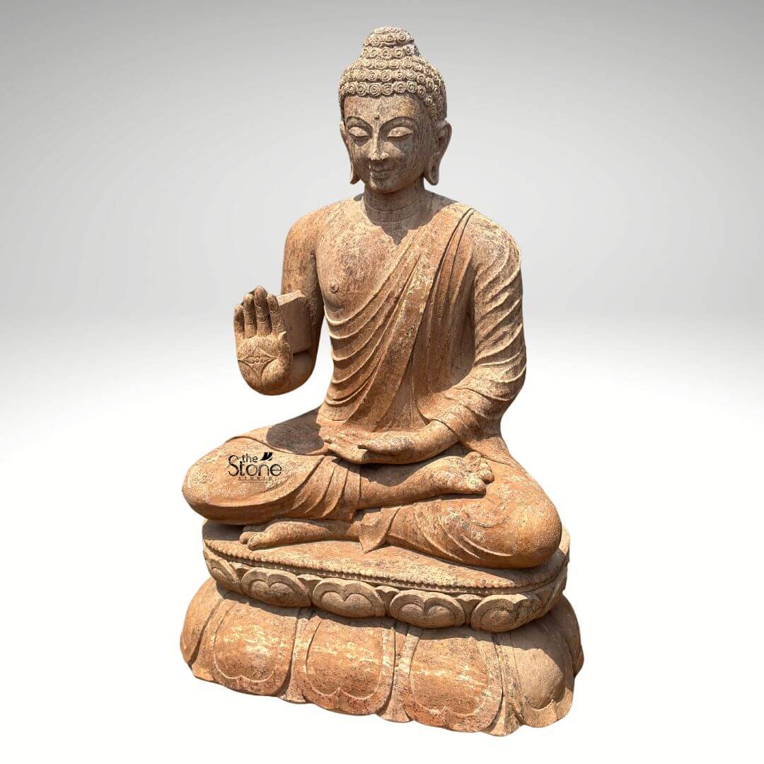 Sandstone Buddha Statue in Abhaya Mudra 3ft 6 A magnificent 3-foot Sitting Buddha Stone Statue stands prominently against a clean light grey background radiating peace and reassurance. The statue carved from a warm reddish-brown sandstone depicts Buddha in a serene meditative posture specifically the full lotus position symbolizing deep concentration and spiritual enlightenment. This classic sitting pose is central to its representation. His right hand is raised to shoulder height with the palm facing outwards fingers extended upwards forming the Abhaya Mudra the gesture of fearlessnes blessing and protection. His left hand rests gently in his lap palm upwards signifying contemplation and receptivity. The natural earthy hue and weathered texture of the sandstone give the sculpture an authentic ancient fee highlighting its intricate details. The Buddha's facial features are exquisitely carved radiating profound peace and wisdom. His eyes are gently closed or cast downwards in contemplation and a subtle serene smile graces his lips. His head is adorned with tightly coiled hair culminating in the ushnisha the cranial bump symbolizing his supreme spiritual knowledge and enlightenment. The flowing drapery of his robes is sculpted with soft elegant folds emphasizing the texture of the sandstone and contributing to the statue's graceful and tranquil form. The rustic worn appearance of the sandstone adds to its charm and gives it a timeless quality suggesting enduring serenity. The Buddha is seated upon a beautifully sculpted double-lotus pedestal with each petal intricately carved symbolizing purity spiritual growth and the emergence of enlightenment from the mundane. The robust and detailed base supports the statue with dignity making it a focal point. This 3-foot Sandstone Buddha statue with its clear depiction of the Abhaya Mudra and its iconic sitting posture is an exquisite example of religious artistry designed to bring blessings peace and aesthetic beauty to any indoor or outdoor space perfectly aligning with an article focusing on Sitting Buddha Stone Statues.