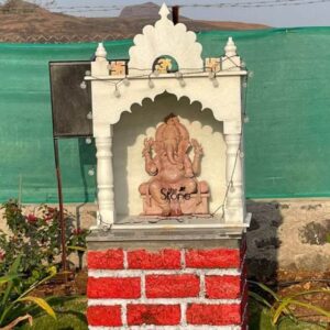 A medium shot captures a beautifully crafted eighteen-inch-tall statue of Lord Ganesha hand-carved from a warm pink sandstone enshrined outdoors. The statue is seated in a traditional graceful posture on a pedestal within a miniature ornate white stone temple structure or mandir which features classic architectural elements like pillars and a decorative shikara top. The Ganesha figure is depicted with four arms holding symbolic attributes and its trunk is curled gracefully to the left. The pink stone exhibits a fine natural granular texture with delicate shallow-relief carvings defining the deity's jewelry crown and the folds of its garments. The entire shrine is mounted securely atop a sturdy square pedestal base constructed from vibrant rough-hewn red bricks which are accented with a clean white painted band at the bottom. This combination of the traditional white temple and rustic red brick base creates a charming contrast making it a prominent focal point. The shrine is set outdoors against a practical and common backdrop in the Pimpri Chinchwad area: a tall solid green mesh fence that separates the property with a glimpse of dry hilly terrain under a clear blue sky visible in the far distance conveying a sense of the local environment. The immediate surroundings are well-kept with patches of green grass and various small garden plants including a flowering bush to the left framing the base of the shrine. A string of warm spherical LED fairy lights is subtly draped around the white temple structure adding an inviting glow. The natural daylight illuminates the scene from the upper right casting soft defined shadows that emphasize the three-dimensional volume of the Ganesha statue and the intricate details of its carving. The composition captures this beloved small Ganesha statue as a cherished home temple piece reflecting a blend of traditional spirituality and contemporary outdoor decor common in Maharashtra.