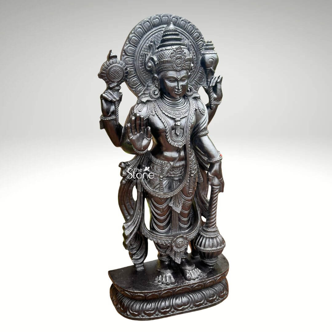 Black Stone Narayana Swamy Statue 3.5ft 10 A detailed full-body photograph captures a majestic forty-one-inch-tall statue of Lord Narayana meticulously hand-carved from a single solid block of deep black stone presenting a commanding devotional figure. The deity is depicted in a traditional standing posture upon an intricately carved circular lotus pedestal which features detailed petal layers and a decorative base. Narayana is shown with four arms radiating divine grace and power making it an ideal sacred art piece for a home temple or place of worship. The statue exhibits a rich dark matte-to-satin finish characteristic of high-quality natural black stone which absorbs light and highlights the depth and complexity of the carvings. The four-armed form of Lord Narayana holds traditional symbolic attributes each intricately defined against the dark textured stone. His upper right hand holds the chakra while his upper left hand holds the shankha. His lower right hand is shown in the abhaya mudra a gesture of fearlessness and blessing and his lower left hand rests gracefully holding the gada. The deity is adorned with elaborate shallow-relief carvings of traditional South Indian temple jewelry including multiple necklaces armlets bracelets a sacred thread and a decorative mukuta all rendered with exceptional precision against the smooth stone. The statue conveys a sense of profound spiritual calm and majesty with the intricate details of his flowing dhoti with elaborate folds the detailed garlands and the serene facial expression all contributing to its devotional appeal. A grand intricately carved prabhavali forms a halo behind the deity adorned with stylized floral and geometric patterns that frame his head and shoulders. The statue is presented against a clean neutral graduated grey background ensuring the focus remains entirely on the detailed figure of Narayana and its artistic craftsmanship perfectly illustrating its suitability as a devotional focal point. The natural stone material ensures durability and a timeless quality.