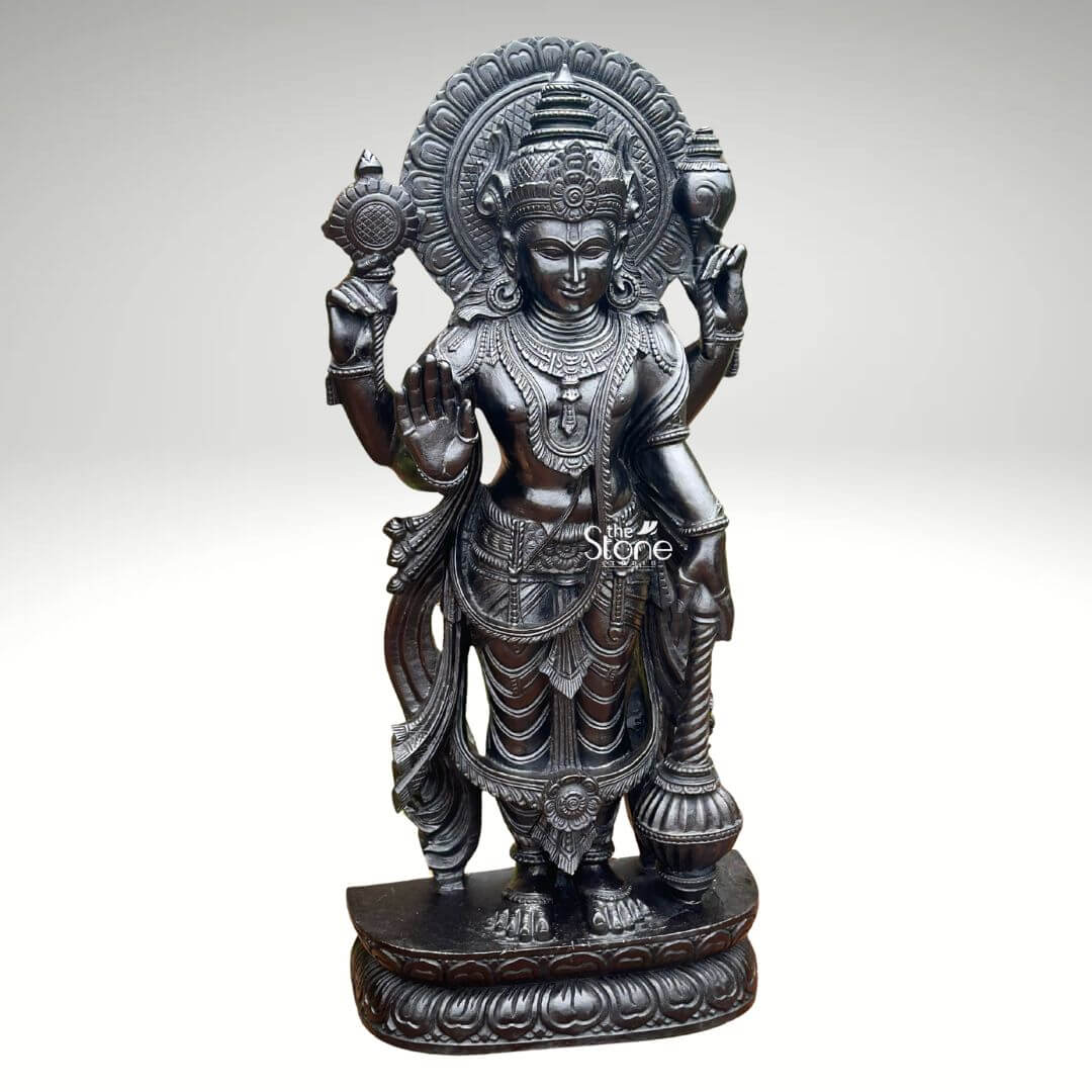 Black Stone Narayana Swamy Statue 3.5ft 11 A detailed full-body photograph captures a majestic forty-one-inch-tall statue of Lord Narayana Swamy meticulously hand-carved from a single solid block of deep black stone presenting a commanding devotional figure. The deity is depicted in a traditional standing posture upon an intricately carved circular lotus pedestal which features detailed petal layers and a decorative base. Narayana is shown with four arms radiating divine grace and power making it an ideal sacred art piece for a home temple or place of worship. The statue exhibits a rich dark matte-to-satin finish characteristic of high-quality natural black stone which absorbs light and highlights the depth and complexity of the carvings. The four-armed form of Lord Narayana holds traditional symbolic attributes each intricately defined against the dark textured stone. His upper right hand holds the chakra while his upper left hand holds the shankha. His lower right hand is shown in the abhaya mudr a gesture of fearlessness and blessing and his lower left hand rests gracefully holding the gada. The deity is adorned with elaborate shallow-relief carvings of traditional South Indian temple jewelry including multiple necklaces armlets, bracelets, a sacred thread and a decorative mukuta all rendered with exceptional precision against the smooth stone. The statue conveys a sense of profound spiritual calm and majesty, with the intricate details of his flowing dhoti with elaborate folds the detailed garlands and the serene facial expression all contributing to its devotional appeal. A grand intricately carved prabhavali forms a halo behind the deity adorned with stylized floral and geometric patterns that frame his head and shoulders. The statue is presented against a clean neutral graduated grey background ensuring the focus remains entirely on the detailed figure of Narayana Swamy and its artistic craftsmanship perfectly illustrating its suitability as a devotional focal point. The natural stone material ensures durability and a timeless quality.