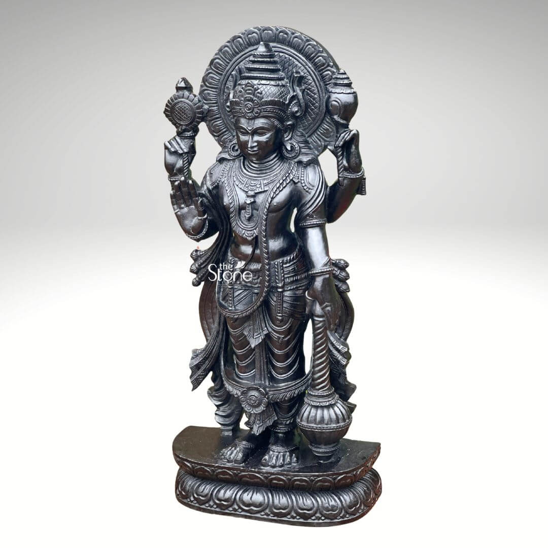 Black Stone Narayana Swamy Statue 3.5ft 12 A detailed full-body photograph captures a majestic forty-one-inch-tall statue of Lord Narayana Swamy meticulously hand-carved from a single solid block of deep black stone presenting a commanding devotional figure. The deity is depicted in a traditional standing posture upon an intricately carved circular lotus pedestal which features detailed petal layers and a decorative base. Narayana is shown with four arms radiating divine grace and power making it an ideal sacred art piece for a home temple or place of worship. The statue exhibits a rich dark matte-to-satin finish characteristic of high-quality natural black stone which absorbs light and highlights the depth and complexity of the carvings. The four-armed form of Lord Narayana holds traditional symbolic attributes each intricately defined against the dark textured stone. His upper right hand holds the chakra while his upper left hand holds the shankha. His lower right hand is shown in the abhaya mudr a gesture of fearlessness and blessing and his lower left hand rests gracefully holding the gada. The deity is adorned with elaborate shallow-relief carvings of traditional South Indian temple jewelry including multiple necklaces armlets bracelets a sacred thread and a decorative mukuta all rendered with exceptional precision against the smooth stone. The statue conveys a sense of profound spiritual calm and majesty with the intricate details of his flowing dhoti with elaborate folds the detailed garlands and the serene facial expression all contributing to its devotional appeal. A grand intricately carved prabhavali forms a halo behind the deity adorned with stylized floral and geometric patterns that frame his head and shoulders. The statue is presented against a clean neutral graduated grey background ensuring the focus remains entirely on the detailed figure of Narayana Swamy and its artistic craftsmanship perfectly illustrating its suitability as a devotional focal point. The natural stone material ensures durability and a timeless quality.