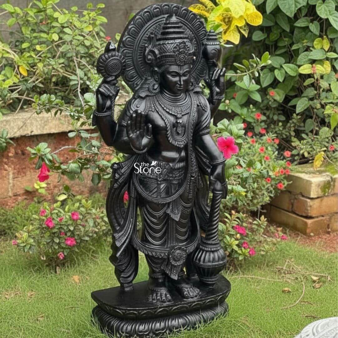 Black Stone Narayana Swamy Statue 3.5ft 3 A majestic full-body photograph captures a four-foot-tall hand-carved statue of Lord Narayana Swamy sculpted from a solid block of deep black stone. The deity is depicted in a traditional standing posture on an intricately carved circular lotus pedestal radiating divine grace and spiritual authority making it an ideal devotional centerpiece for a home temple or place of worship. The statue exhibits a rich dark matte finish characteristic of high-quality natural black stone which absorbs light and emphasizes the depth and complexity of the carvings. The four-armed form of Lord Narayana holds traditional symbolic attributes each meticulously defined against the dark stone. His upper right hand holds the chakra while his upper left hand holds the shankha. His lower right hand is shown in the abhaya mudra a gesture of fearlessness and blessing and his lower left hand rests gracefully holding the gada. The deity is adorned with elaborate shallow-relief carvings of traditional South Indian temple jewelry including multiple necklaces armlets bracelets a sacred thread and a decorative mukuta all rendered with exceptional precision. The statue conveys a sense of profound spiritual calm and majesty with the intricate details of his flowing dhoti with elaborate folds the detailed garlands and the serene facial expression all contributing to its devotional appeal. A grand intricately carved prabhavali forms a halo behind the deity adorned with stylized floral and geometric patterns that frame his head and shoulders. The statue is presented outdoors in a lush sunlit garden setting standing on a patch of green grass with various flowering plants including vibrant pink and yellow blooms creating a natural sanctuary that complements the sacred figure. The natural daylight illuminates the scene highlighting the rich textures of the hand-carved stone and the peaceful atmosphere of the garden. The natural stone material ensures durability and a timeless quality.