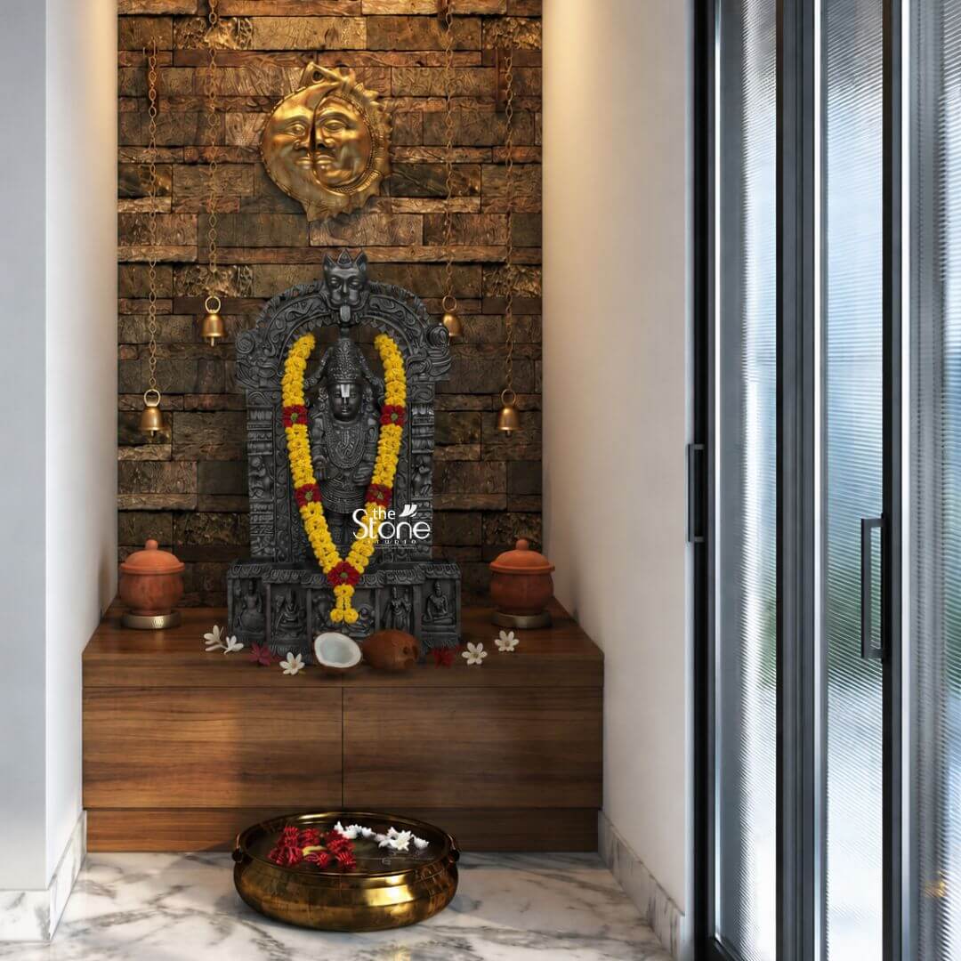 Sri Srinivasa Tirupati Balaji Statue 2.5ft 3 A reverential three-quarter view photograph captures a meticulously hand-carved Sri Srinivasa Swamy stone statue standing 2.5 feet tall within a dedicated home temple niche. The statue is sculpted from a single block of dark dense blackstone polished to a deep matte finish that accentuates the intricate sculptural details. The deity also known as Tirupati Balaji is depicted in a samabhanga posture upon a lotus pedestal which is itself supported by an ornate multi-tiered rectangular base. A vibrant garland of fresh yellow marigolds and red flowers drapes gracefully around the deity's neck contrasting beautifully with the dark stone. The statue is framed by a traditional South Indian prabhavali adorned with floral motifs and culminating in a kirtimukha face at the apex flanked by two smaller brass hanging temple bells.The stone idol is enshrined on a contemporary wooden wall-mounted shelf which serves as the altar. On the shelf flanking the deity are two traditional terracotta pots with lids resting on small brass stands. At the base of the statue are various puja offerings including a halved coconut a whole brown coconut and scattered white flowers like jasmine and frangipani. The entire temple space is backed by a textured dark brown stacked-stone wall which adds a rustic element to the modern interior. Above the archway a large intricately carved golden sun and moon face plaque is mounted, further enhancing the divine atmosphere. The setting is a modern home corner with the temple niche situated next to a floor-to-ceiling glass door with dark frames offering a glimpse of the outdoors. The floor is covered in light grey marble tiles and in the foreground a large ornate brass urli filled with water floating red and white flowers sits as a decorative element. The lighting is warm and focused emanating from hidden LED strips within the niche highlighting the texture of the blackstone and the gold accents. The photograph's clear focus on the statue's form and texture provides a comprehensive view for the viewer making it a perfect visual reference for an article discussing such sacred stone statues.