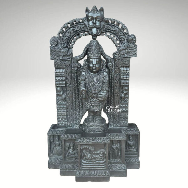 A detailed front-facing photograph presents a magnificent 2.5-foot-tall statue of Sri Srinivasa Tirupati Balaji meticulously hand-carved from a single block of dark dense blackstone. The statue depicts Lord Balaji standing in a samabhanga posture on a lotus pedestal which is itself supported by an ornate multi-tiered rectangular base. The dark stone has a deep matte finish that highlights the intricate sculptural details. Garuda the celestial eagle-mount is prominently carved at the apex of the elaborate prabhavali that frames the main deity. Below Garuda the arch is adorned with traditional floral and scrollwork motifs culminating in makara heads at the base of the pillars. Lord Balaji's divine form is richly adorned with finely carved traditional ornaments including multiple layered necklaces elaborate armbands bracelets and anklets. His forehead bears the distinct Vaishnava U-shaped tilak mark and his eyes are half-closed in a serene benevolent expression. His upper right hand holds a discus while his upper left hand holds a conch shell. His lower right hand is raised in the Varada mudra and his lower left hand rests gracefully on his hip. The fine detail extends to his draped clothing which is textured to resemble silk fabric held by a jeweled girdle. The rectangular base beneath the main figure is a work of art in itself featuring intricate bas-relief panels on its visible front face. The central panel depicts the reclining form of Lord Ranganatha resting on the serpent Adisesha attended by consorts. This central scene is flanked by smaller detailed carvings of sages devotees and other deities within architectural niches. The entire sculpture from the ornate pedestal to the top of the arch reflects the traditional South Indian Hoysala and Vijayanagara architectural styles. This 2.5-foot blackstone Tirupati Balaji statue is an exceptional piece of devotional art perfect for a home shrine or as a central exhibit capturing the spiritual essence and cultural heritage of the Venkateswara Temple. The photograph's clear focus on the statue's form and texture provides a comprehensive view for the viewer.