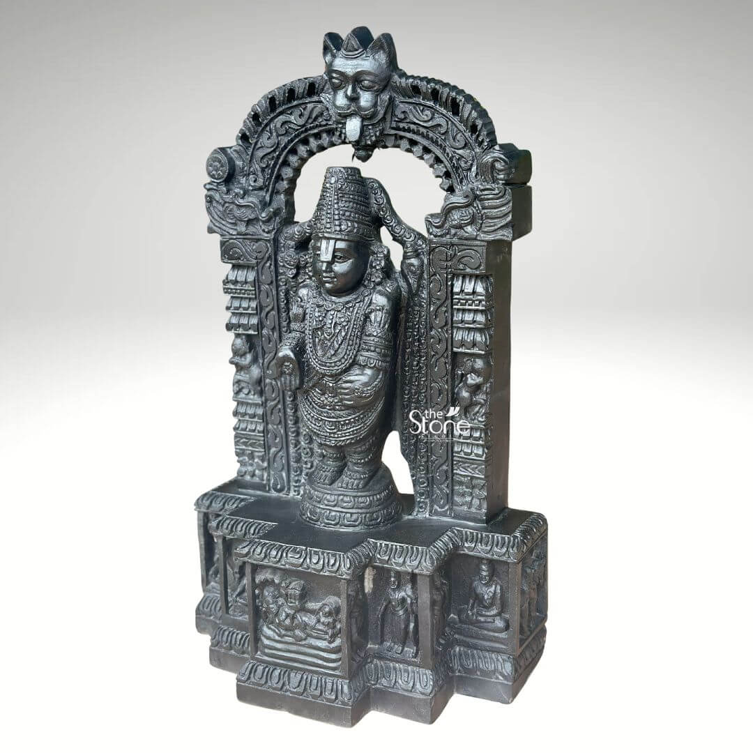 Sri Srinivasa Tirupati Balaji Statue 2.5ft 5 A detailed three-quarter view photograph captures a magnificent 2.5-foot-tall statue of Sri Venkateswara also known as Tirupati Balaji meticulously hand-carved from a single block of dark dense blackstone. The statue depicts Lord Balaji standing in a samabhanga posture on a lotus pedestal, which is itself supported by an ornate multi-tiered rectangular base. The dark stone has a deep matte finish that accentuates the intricate sculptural details. Garuda the celestial eagle-mount is prominently carved at the apex of the elaborate prabhavali that frames the main deity. Below Garuda the arch is adorned with traditional floral and scrollwork motifs culminating in makara heads at the base of the pillars.Lord Balaji's divine form is richly adorned with finely carved traditional ornaments, including multiple layered necklaces elaborate armbands bracelets and anklets. His forehead bears the distinct Vaishnava U-shaped tilak mark and his eyes are half-closed in a serene, benevolent expression. His upper right hand holds a discus while his upper left hand holds a conch shell. His lower right hand is raised in the Varada mudra and his lower left hand rests gracefully on his hip. The fine detail extends to his draped clothing which is textured to resemble silk fabric held by a jeweled girdle. The rectangular base beneath the main figure is a work of art in itself featuring intricate bas-relief panels on its visible front face. The central panel depicts the reclining form of Lord Ranganatha resting on the serpent Adisesha attended by consorts. This central scene is flanked by smaller detailed carvings of sages devotees and other deities within architectural niches. The entire sculpture from the ornate pedestal to the top of the arch reflects the traditional South Indian Hoysala and Vijayanagara architectural styles. This 2.5-foot blackstone statue is an exceptional piece of devotional art perfect for a home shrine or as a central exhibit capturing the spiritual essence and cultural heritage of the Venkateswara Temple. The photograph's clear focus on the statue's form and texture provides a comprehensive view for the viewer.