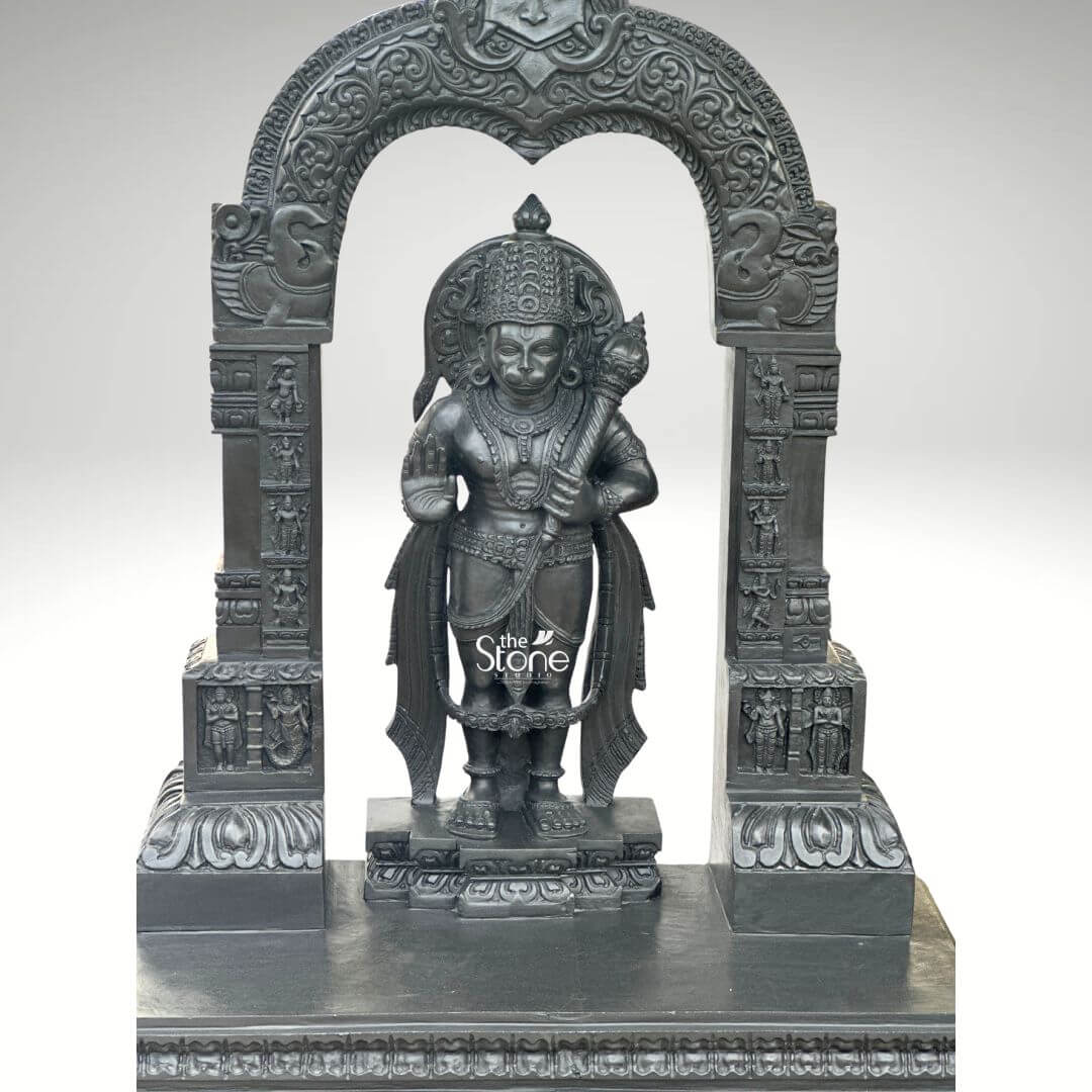 Black Stone Hanuman Statue with Arch 3ft 6 A magnificent 3-foot Standing Bajrangbali Stone Statue intricately carved from deep black stone stands prominently within an ornate archway against a clean light grey background creating a powerful and reverent display. The statue depicts Lord Hanuman also known as Bajrangbali in a traditional standing posture radiating immense strength unwavering devotion and a vigilant readiness for service. He is adorned with an elaborate crown traditional jewelry around his neck chest, and arms and finely sculpted flowing garments all rendered with exceptional detail in the lustrous dark stone. In his left hand he firmly grasps his formidable gada a potent symbol of his immense power and valor while his right hand is raised in a gesture of Abhaya Mudra offering fearlessness and divine protection to all devotees. The craftsmanship of this Blackstone Hanuman statue is truly outstanding with every minute detail meticulously brought to life. His facial features including his serene yet resolute expression are precisely rendered conveying his spiritual resolve. The subtle depiction of his musculature beneath his garments hints at his legendary strength and agility perfectly suiting the epithet Bajrangbali meaning the strong one. Lord Hanuman stands on a multi-tiered black stone pedestal which is seamlessly integrated into the broader base of the entire archway structure providing both stability and enhancing its grand temple-like presence. The statue is dramatically framed by a beautifully carved decorative arch also crafted from black stone. This arch features intricate traditional patterns floral motifs and smaller divine figures or mythological scenes meticulously carved into its supporting pillars and along the curve of the arch. These surrounding architectural elements elevate the spiritual grandeur and cultural richness of the entire piece reminiscent of ancient temple entrances. This impressive 3-foot Standing Bajrangbali Stone Statue with its integrated decorative arch serves as an ideal representation for an article focusing on such a divine sculpture symbolizing unwavering loyalty courage profound spiritual strength and the iconic powerful form of Lord Hanuman.