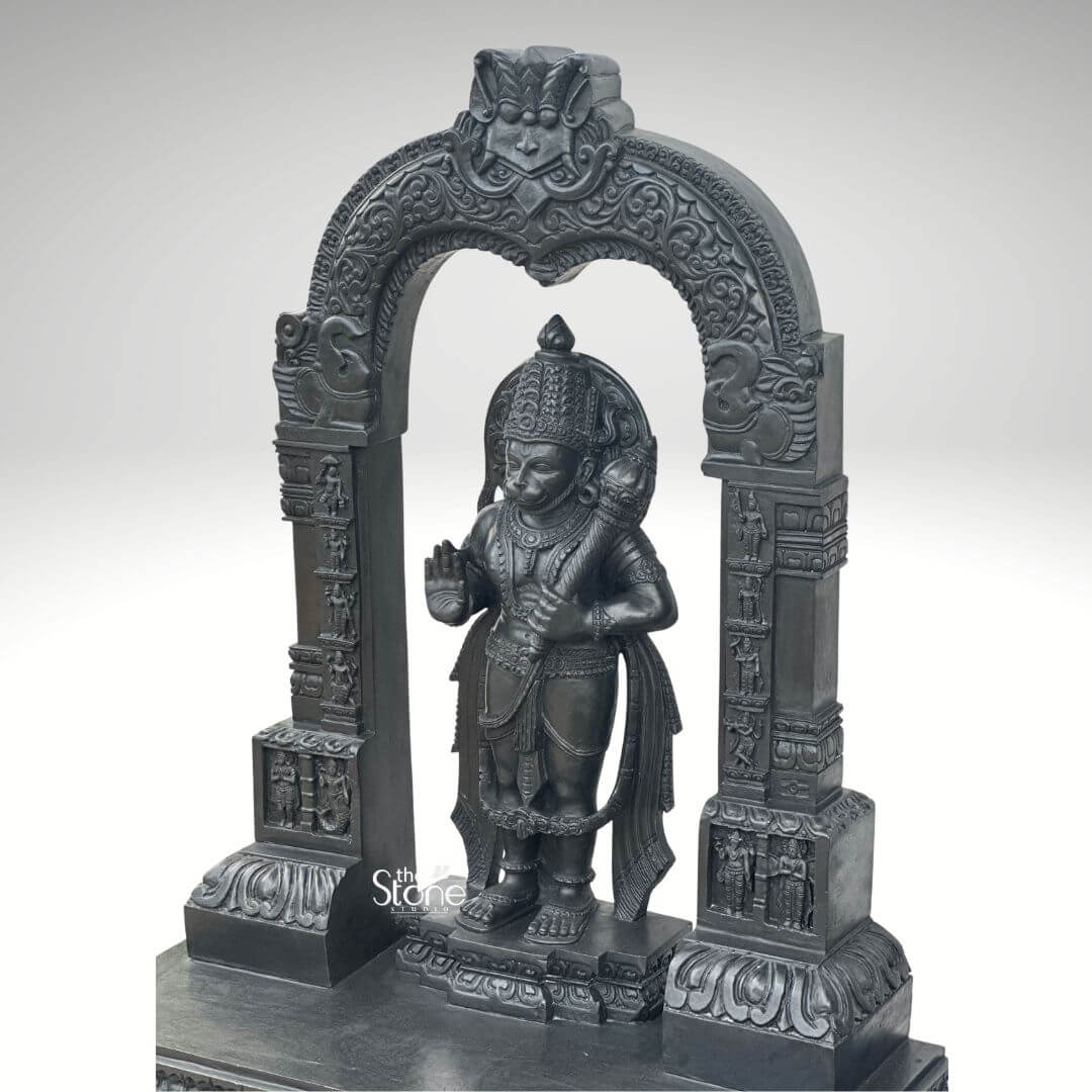 Black Stone Hanuman Statue with Arch 3ft 7 A magnificent 3-foot Standing Hanuman Statue in Black Stone is presented with impressive detail within an ornate archway set against a clean light grey background creating a powerful and reverent display. The statue depicts Lord Hanuman in a traditional standing posture radiating immense strength unwavering devotion and a vigilant readiness for service. He is intricately carved from deep lustrous black stone with an elaborate crown traditional jewelry adorning his neck chest, and arms and finely sculpted flowing garments all rendered with exceptional precision. In his left hand he firmly grasps his formidable gada a potent symbol of his immense power and valor while his right hand is raised in a gesture of Abhaya Mudra offering fearlessness and divine protection to all devotees. The craftsmanship of this Blackstone Hanuman statue is truly outstanding with every minute detail meticulously brought to life. His facial features including his serene yet resolute expression are precisely rendered conveying his spiritual resolve and powerful persona. The subtle depiction of his musculature beneath his garments hints at his legendary strength and agility. Lord Hanuman stands on a multi-tiered black stone pedestal which is seamlessly integrated into the broader base of the entire archway structure providing both stability and enhancing its grand temple-like presence perfectly suited for a standing deity. The statue is dramatically framed by a beautifully carved decorative arch also crafted from black stone. This arch features intricate traditional patterns floral motifs and smaller divine figures or mythological scenes meticulously carved into its supporting pillars and along the curve of the arch. These surrounding architectural elements elevate the spiritual grandeur and cultural richness of the entire piece reminiscent of ancient temple entrances. This impressive 3-foot Standing Hanuman Statue in Black Stone with its integrated decorative arch serves as an ideal representation for an article focusing on such a divine sculpture embodying unwavering loyalty courage profound spiritual strength and exceptional artistry.
