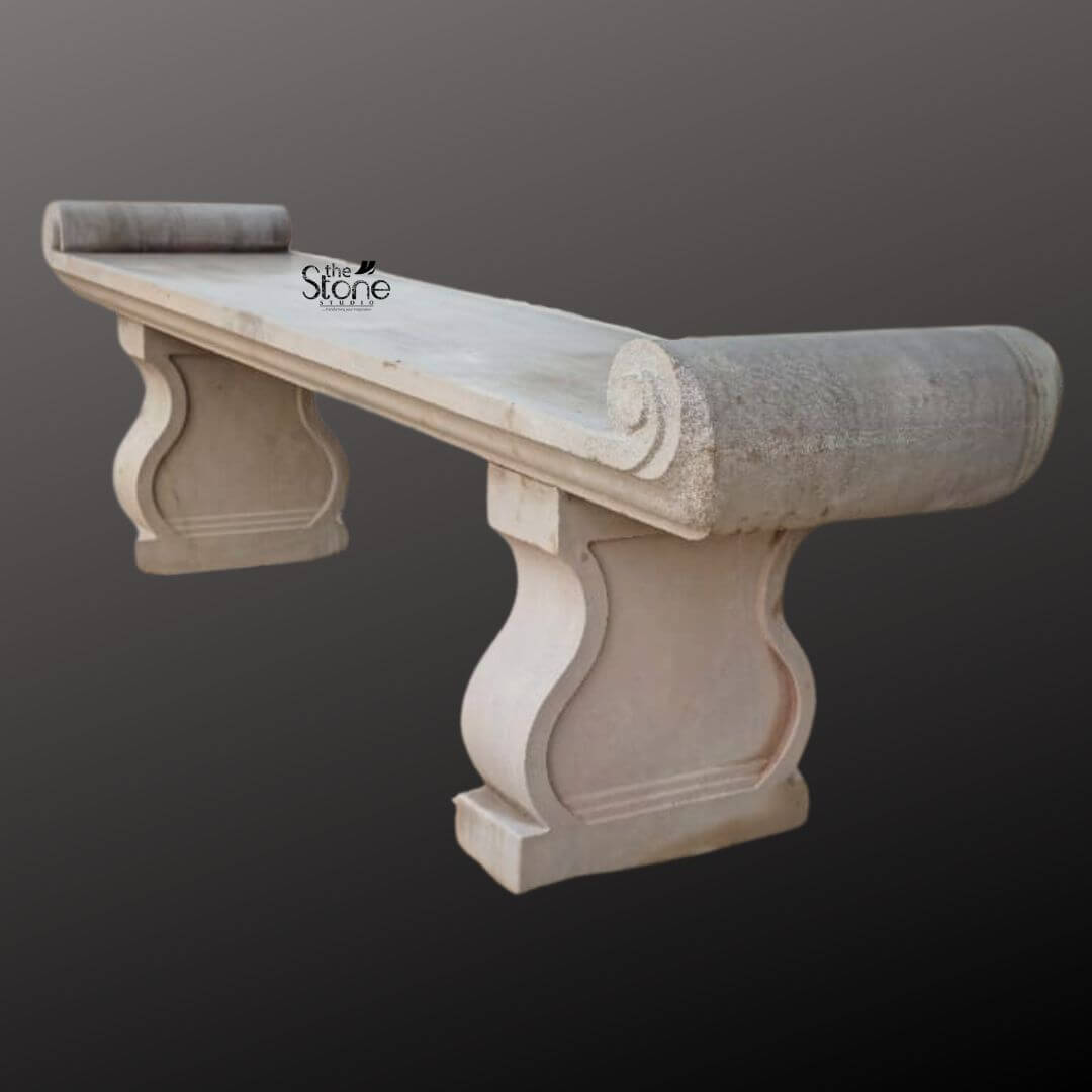 Sandstone Garden Bench 6ft 6 A six-foot-long sandstone garden bench stands against a seamless dark grey studio background showcasing its elegant classical design. The bench is hand-carved from natural light beige sandstone exhibiting a beautifully textured porous surface that adds an authentic rustic charm. Its long flat seat is supported by two substantial decoratively sculpted legs. These legs feature a graceful S-curve or hourglass profile resting on sturdy block-like bases that anchor the entire piece. The craftsmanship is evident in the smooth yet hand-hewn finish of the stone which catches the light and highlights its natural variations in color and texture from pale cream to subtle warm brown tones. The design of this sandstone bench is distinctly traditional inspired by historical European garden furniture. The seat itself is not plain it terminates at both the left and right ends with large perfectly formed scrolled volutes or rolled ends. These sculptural elements not only provide comfortable armrests but also define the bench's aesthetic lending it a sophisticated and timeless appeal. The overall form is balanced and substantial suggesting durability and a permanent place within a garden landscape. The stone appears heavy and solid capable of withstanding the elements for decades. This six-foot sandstone bench is the perfect embodiment of a functional and decorative Stone Bench for Garden. Its generous length provides ample seating for multiple people while its robust stone construction makes it an ideal choice for outdoor settings like patios courtyards or nestled within garden pathways. The light neutral color of the sandstone harmonizes beautifully with surrounding greenery flowers and other natural elements. This bench is not just a piece of furniture it is a sculptural addition to a garden designed to offer a comfortable and contemplative spot to rest and enjoy the beauty of the outdoors.