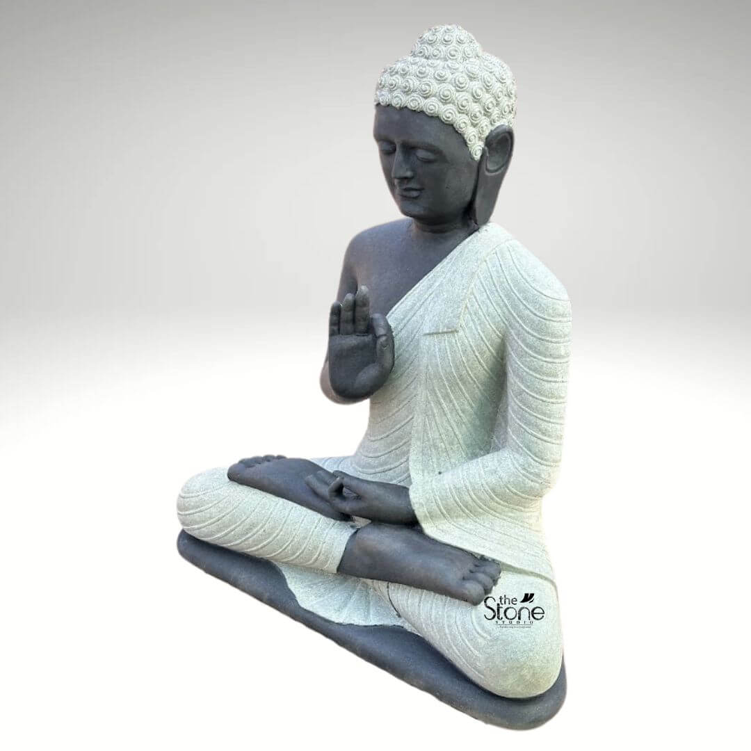 Two Tone Buddha Stone Statue for Home 3ft 7 A serene, hand-carved Blackstone Buddha statue sits in a traditional meditative pose, presented in a clean studio setting against a soft grey background. The three-foot-tall statue showcases a striking two-tone finish, with the Buddha's body and head carved from a deep, dark blackstone, exhibiting a smooth, polished texture that highlights the rich, dark tones. This contrasts beautifully with his monastic robes, which are rendered in a contrasting lighter grey or off-white stone, featuring a subtle, granular texture. This distinct separation emphasizes the form of the robes as they drape over his crossed legs and shoulders, adding depth and visual interest to the sacred art. The Buddha is depicted seated with his legs crossed in a full lotus position (padmasana) on a simple, integrated dark blackstone base. His hands are gracefully positioned in the vitarka mudra, symbolizing discussion and the transmission of teachings, with the right hand raised and the thumb and index finger forming a circle, while the left hand rests in his lap. His facial expression is calm and contemplative, with downcast eyes, a gentle smile, and tightly curled hair leading to a cranial ushnisha, all rendered with precise craftsmanship that honors traditional iconography. The overall composition evokes a sense of peace, tranquility, and spiritual harmony. This three-foot Blackstone Buddha statue is an ideal illustration for an article discussing a Stone Buddha for Home & Garden, demonstrating how such a piece can transform an indoor or outdoor space into a peaceful sanctuary. Its substantial size ensures it functions as a commanding focal point in a living room, entryway, or garden nook, while the durable blackstone construction ensures it is both a beautiful and a meaningful addition to the home environment. The traditional design brings a sense of sacred history and calm to any setting, whether nestled within a lush garden or placed as a spiritual anchor in an interior. The detailed carving and harmonious integration of elements highlight its value as a piece of sacred art.