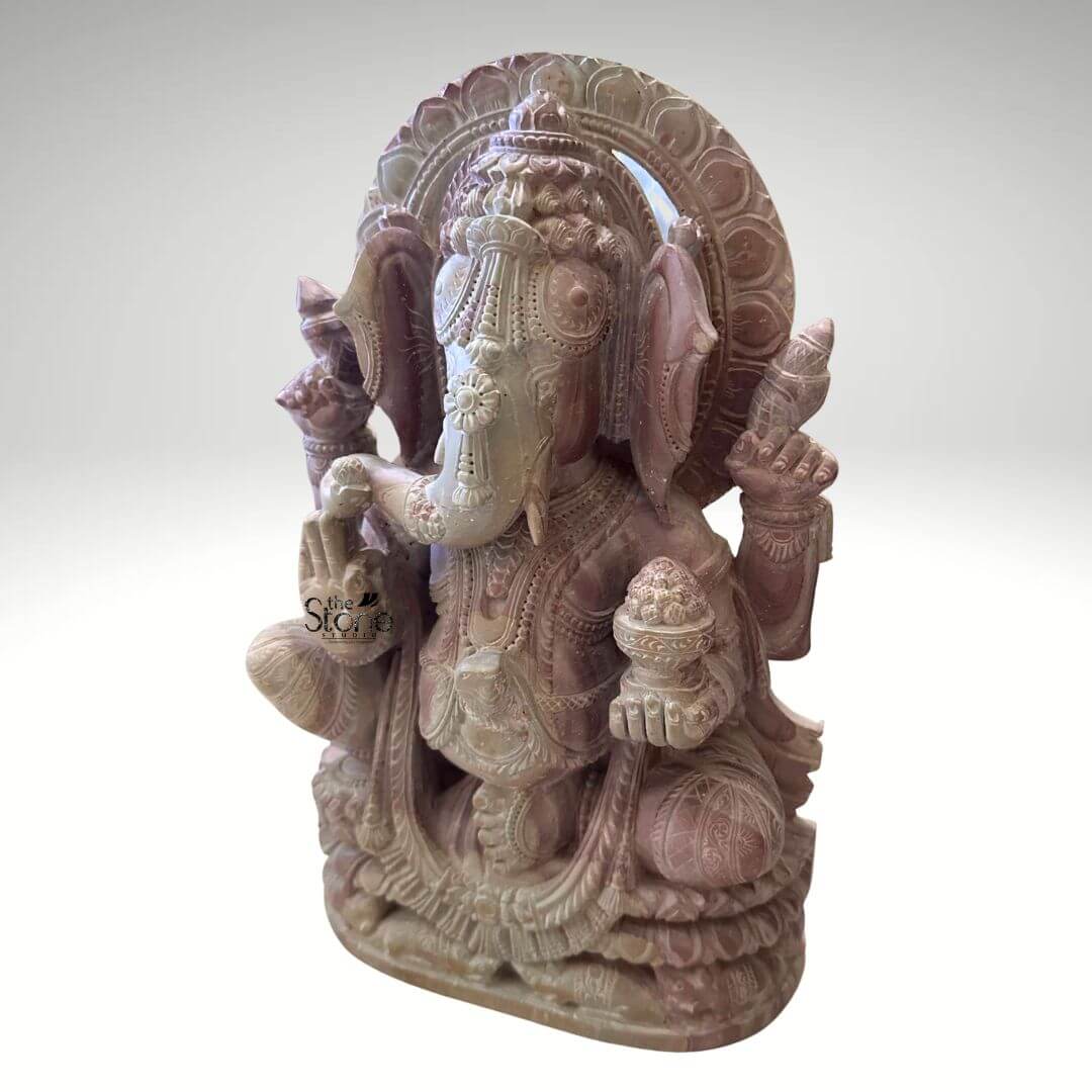 Pink Stone Ganpati Idol for Home 12" 4 A three-quarter front view photograph captures a hand-carved stone Ganesha statue measuring twelve inches in height sculpted from a natural pink stone. The statue depicts the revered elephant-headed Hindu deity in a seated meditative pose with one leg gently folded over the other resting upon a multi-tiered lotus-carved pedestal base. The pink stone exhibits a soft variegated hue ranging from pale rose to deeper mauve with subtle natural veining and crystalline inclusions throughout giving the idol a warm and earthy texture. The entire sculpture is highly detailed with intricate carvings covering every aspect of the divine form.Ganesha is adorned with traditional deeply carved jewelry, including elaborate layered necklaces armbands anklets and a richly patterned crown that frames his serene face. His eyes are half-closed in contemplation and his trunk curves gracefully to his left holding a single perfectly formed modak. The four-armed deity holds symbolic attributes in his hands, all rendered with fine precision: his upper right hand holds a goad his upper left hand holds a noose his lower right hand is raised in the Abhaya mudra and his lower left hand holds a bowl overflowing with more modaks. A decorative prabhavali with intricate floral and geometric motifs rises behind his head completing the divine iconography. This twelve-inch pink stone Ganesha statue is an ideal choice for a home temple or as a sacred centerpiece in a living space. Its compact size indicates its suitability for placement on a home altar a prominent shelf or within a dedicated puja room, bringing a sense of spiritual tranquility and cultural artistry to any interior. The choice of pink stone adds a gentle inviting color to the decor while the detailed craftsmanship reflects a deep respect for traditional iconography. The photograph's clear focus on the statue's texture form and craftsmanship provides a comprehensive view for the viewer making it a perfect visual reference for an article on selecting a Ganesha statue for a home temple