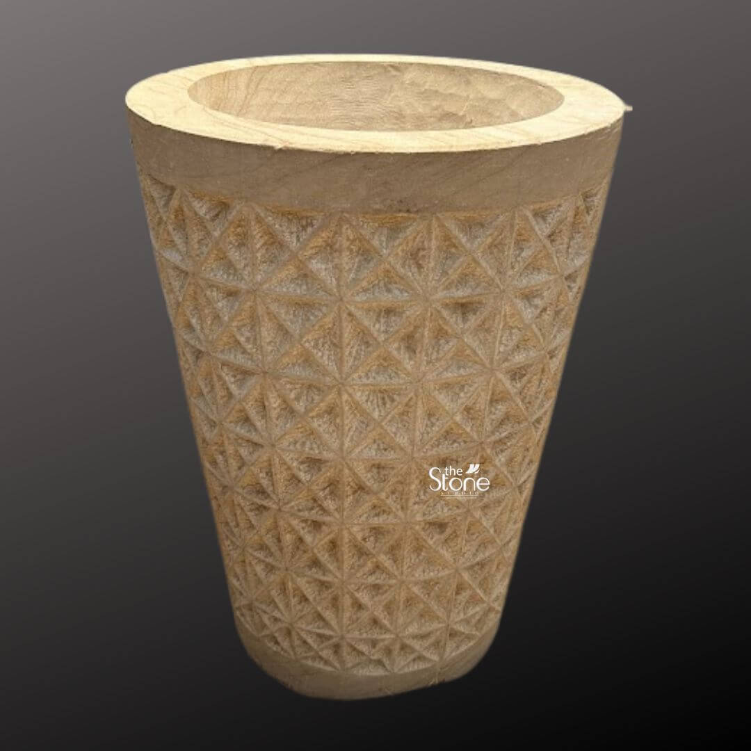Natural Sandstone Planter Pot 2ft 2 A full-view photograph of a magnificent 2-foot tall stone garden planter pot presented against a dark neutral gray gradient background that accentuates its form. The planter is hand-carved from a solid block of warm textured beige sandstone showcasing a rich natural grain and tonal variations that highlight its geological origin. It is fashioned into a classic slightly tapered cylindrical shape providing a robust and stable structure for large plants. The overall impression is one of rustic elegance and durable craftsmanship perfectly suited for enhancing any garden or outdoor area. The central body of the stone garden planter pot is adorned with an intricate deeply incised geometric pattern. This pattern consists of a repeating tessellation of diamond and triangular facets creating a textured three-dimensional surface that catches the light and adds significant visual depth. The precision and detail of the carving demonstrate a high level of artisanal skill turning the functional object into a piece of sculptural art. The natural porosity of the sandstone is clearly visible emphasizing its suitability for outdoor environments where it can withstand the elements while allowing soil and plants to breathe. This substantial 2-foot stone planter pot is a versatile and attractive choice for any landscaping project. Its scale and intricate design make it an ideal focal point for a patio deck or garden path providing a sturdy and stylish home for large flowering plants or small shrubs. The image clearly conveys the texture scale and elaborate geometric carving of the planter making it a perfect visual reference for an article discussing natural stone elements in garden design and the beauty of hand-carved sandstone products.