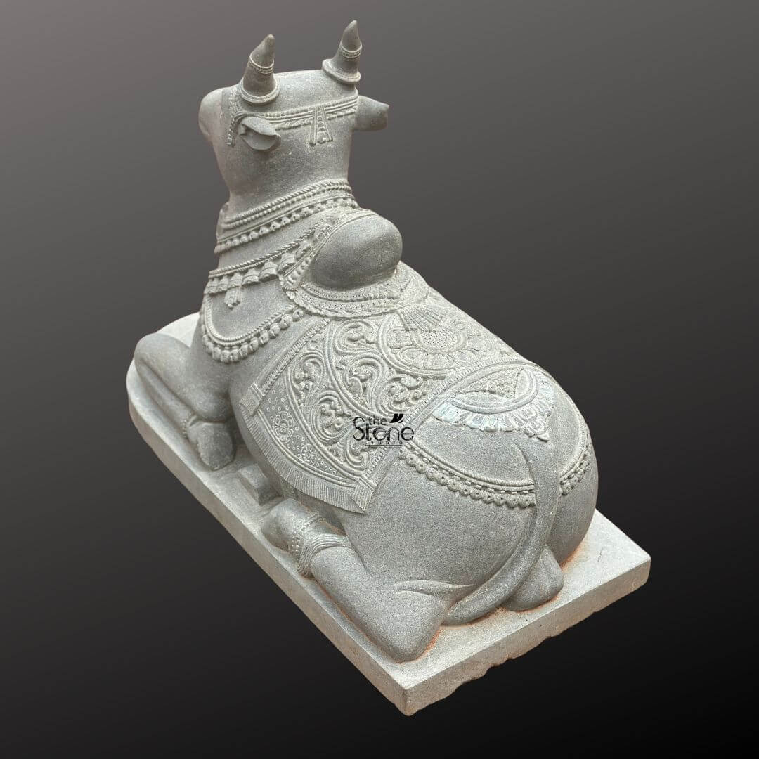 Nandikeshwara Stone Statue 45" 9 A high-angle three-quarter studio photograph showcases a meticulously hand-carved blackstone Nandi bull statue presented in a traditional recumbent pose. The sacred idol is positioned on a plain elevated rectangular stone plinth set against a smooth dark grey gradient background that emphasizes its form. The statue exhibits the dark rich and matte finish characteristic of premium blackstone meticulously worked to create both smooth surfaces and deep-relief textures. Nandi is depicted resting on its folded legs with its head turned gently to the viewer's left conveying a sense of peaceful devotion. The entire body of the Nandi statue is richly adorned with elaborate deeply carved ornamentation that highlights the artisan's skill. Its head features detailed eyes ears and stylized horns, encircled by a delicate headpiece. Multiple layers of finely carved beaded necklaces and chains drape around its neck with the texture of the stone mimicking heavy jewelry. The sculpture conveys a sense of deep spiritual reverence through its intricate craftsmanship, befitting its role as a central temple deity. The body of the Nandi is covered by a beautifully patterned saddle blanket that drapes over its back and sides. The blanket is adorned with complex deep-relief floral motifs geometric patterns and a textured border, finished with detailed fringes along the lower edge. Below the blanket an intricately carved girth strap secures it ensuring the sculpture embodies the rich ceremonial decoration required for temple iconography. The overall impression is one of ancient tradition and masterful artistry making it a fitting subject for articles discussing sacred temple sculpture.