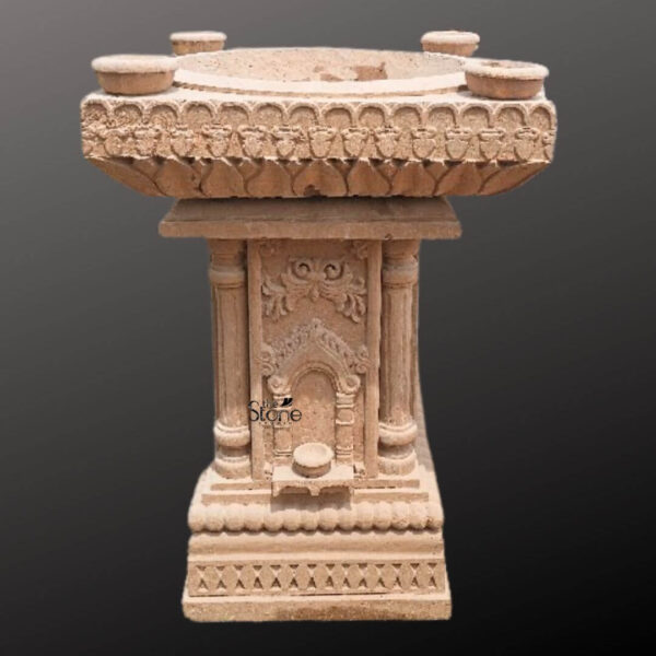 A detailed studio photograph presents a magnificent hand-carved brown sandstone Tulsi planter designed in the traditional temple style. The planter rests on a square base with a decorative geometric lattice pattern supporting a central column. The column features four fluted pilasters and an arched niche on the front face which contains a small carved pedestal for a traditional oil lamp. Above the niche elaborate floral and scrollwork motifs adorn the pillar showcasing the artisan's skillful craftsmanship. The natural warm granular texture of the brown sandstone is visible across the entire structure. The planter culminates in a large square-shaped bowl that serves as the main planting bed for the sacred Tulsi plant. The exterior rim of the bowl is richly decorated with a continuous band of deeply carved lotus petals a motif signifying purity and divinity in Indian culture. At each of the four corners of the bowl's rim there is a smaller integrated circular platform designed to hold traditional oil lamps or small offerings during worship. This thoughtful design integrates the practical need for a planter with the sacred function of a home shrine. This brown sandstone Tulsi planter is an ideal piece of spiritual decor for a home embodying ancient tradition and masterful artistry. Its presence in a living space balcony or garden creates a sacred focal point perfect for the daily worship of Goddess Tulsi. The durable sandstone material ensures that it can be placed either indoors or outdoors where the natural weathering will only enhance the beauty of its intricate carvings over time. This planter beautifully merges cultural significance with aesthetic appeal making it a cherished addition to any home.