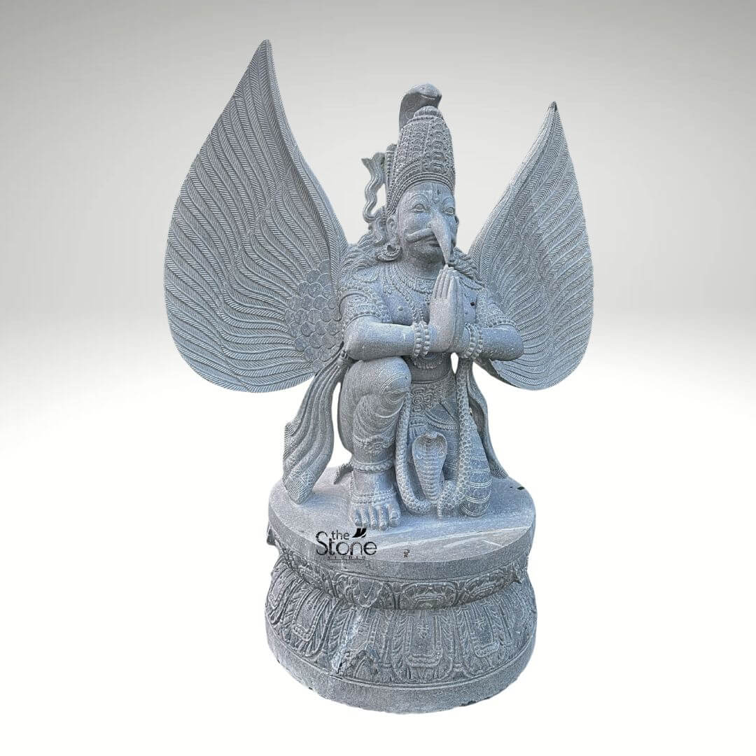 Grey Stone Garuda Statue 5.5ft 9 A full-length front-facing photograph captures a magnificent 5.5-foot-tall Temple Garuda Statue meticulously hand-carved from a single block of natural grey stone. The statue presents the mythical bird-deity Garuda in al reverent kneeling posture positioned upon a robust circular lotus-carved pedestal base. His hands are pressed together in the Anjali Mudra a gesture of prayer and devotion held near his chest. Garuda has a powerfu human-like torso with an eagle-like face defined by a prominent beak, piercing eyes and a traditional crown with intricate, detailed patterns. His body is adorned with finely carved traditional ornaments including layered necklaces armbands bracelets and a sacred thread all contributing to his divine appearance. A defining feature of this temple Garuda statue is its massive fully spread wings which rise dramatically behind him mirroring his height. Each wing feather is individually and delicately carved creating a texture that is both visually detailed and structurally impressive, conveying a sense of power and spiritual elevation. The grey stone itself possesses a natural slightly matte finish with subtle crystalline variations giving the sculpture an authentic ancient feel appropriate for a temple setting and underscoring the traditional craftsmanship. A coiled serpent is visibly carved beneath one of his feet further enhancing the traditional iconography associated with this celestial mount of Lord Vishnu. This five-and-a-half-foot grey stone statue is an exemplary piece of traditional Indian temple art making it a perfect subject for an article discussing Garuda statues in a religious context. Its commanding size and detailed craftsmanship make it suitable for placement within a temple mandapam a shrine or as an architectural element. The choice of durable grey stone ensures its longevity making it a timeless investment for spiritual and aesthetic appreciation. The image clearly displays the unique iconographic elements and the high level of artistry that define such a sacred stone idol.