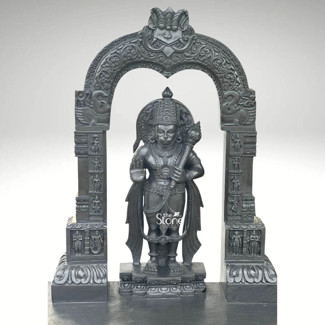 Black Stone Hanuman Statue with Arch 3ft 8 A magnificent 3-foot Temple Hanuman Statue in Black Stone, intricately carved, stands majestically within a grand ornate archway presented against a clean light grey background evoking the solemnity of a sacred temple. The statue depicts Lord Hanuman in a traditional standing posture exuding immense strength unwavering devotion and a vigilant readiness for service. He is richly adorned with an elaborate crown traditional jewelry around his neck chest and arms and finely sculpted flowing garments all rendered with exceptional detail in the lustrous deep black stone. In his left hand he firmly grasps his formidable gada a potent symbol of his immense power and valor while his right hand is raised in a gesture of Abhaya Mudra offering fearlessness and divine protection to all devotees. The craftsmanship of this Blackstone Hanuman statue is truly outstanding with every minute detail meticulously brought to life suitable for a revered temple setting. His facial features including his serene yet resolute expression are precisely rendered conveying his spiritual resolve and powerful persona. The subtle depiction of his musculature beneath his garments hints at his legendary strength and agility. Lord Hanuman stands on a multi-tiered black stone pedestal which is seamlessly integrated into the broader base of the entire archway structure providing both stability and enhancing its grand temple-like presence. The statue is dramatically framed by a beautifully carved decorative arch also crafted from black stone. This arch features intricate traditional patterns elaborate floral motifs and smaller divine figures or mythological scenes meticulously carved into its supporting pillars and along the curve of the arch. These surrounding architectural elements elevate the spiritual grandeur and cultural richness of the entire piece directly reminiscent of ancient temple entrances and mandapams. This impressive 3-foot Temple Hanuman Statue in Black Stone with its integrated decorative arch serves as an ideal representation for an article focusing on such a divine sculpture symbolizing unwavering loyalty courage profound spiritual strength and embodying the essence of a sacred temple deity.