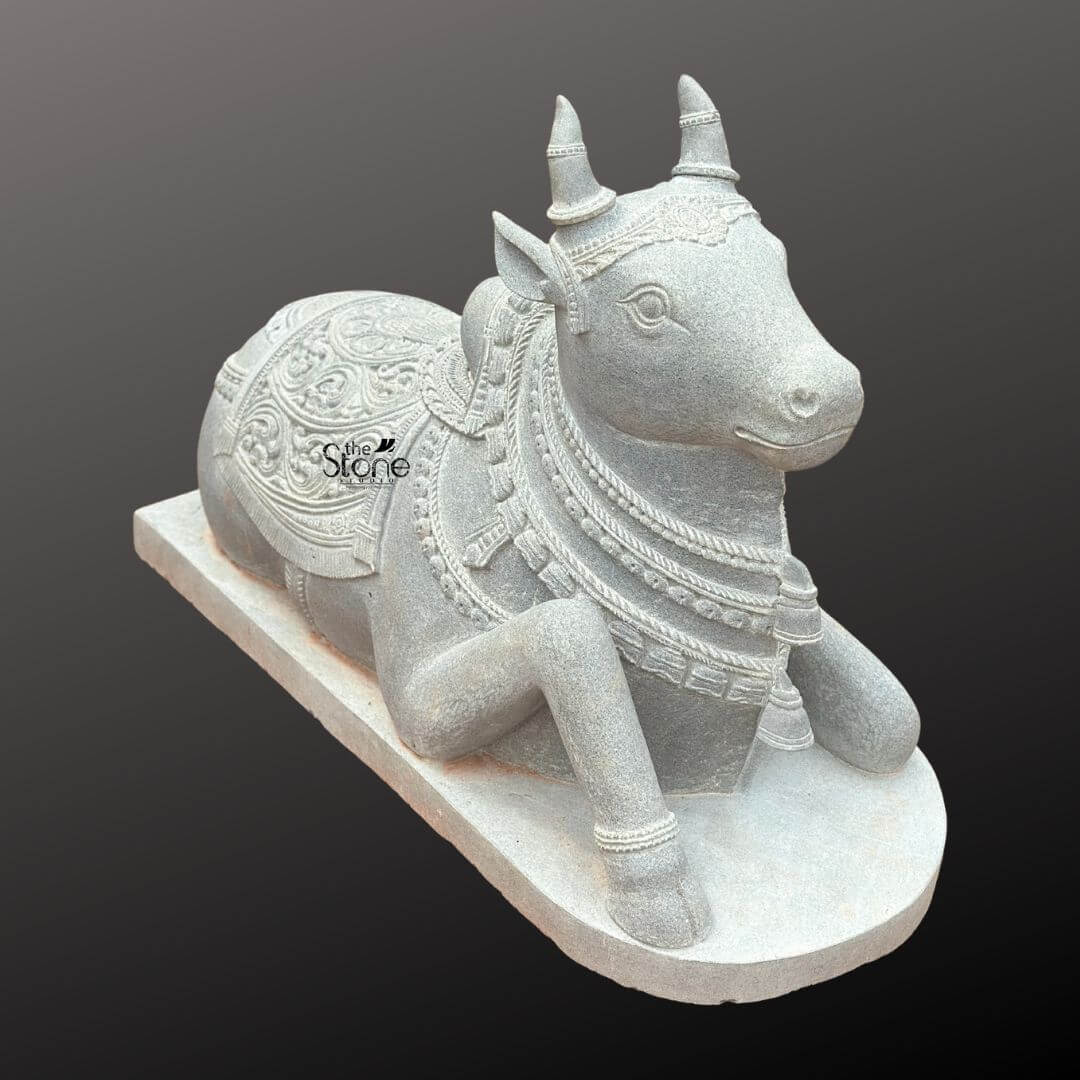 Nandikeshwara Stone Statue 45" 10 A detailed, elevated three-quarter studio photograph showcases a magnificent hand-carved blackstone Nandi bull statue. The sacred idol is presented in a traditional recumbent pose resting on a plain elevated rectangular stone plinth set against a smooth, dark grey gradient background that emphasizes its form. The statue exhibits the dark rich and matte finish characteristic of premium blackstone meticulously worked to create both smooth surfaces and deep-relief textures. Nandi is depicted resting on its folded legs with its head turned gently to the viewer's left conveying a sense of peaceful devotion. The entire body of the Nandi statue is richly adorned with elaborate deeply carved ornamentation that highlights the artisan's skill. Its head features detailed eyes ears and stylized horns encircled by a delicate headpiece. Multiple layers of finely carved beaded necklaces chains and bell pendants drape around its neck with the texture of the stone mimicking heavy temple jewelry. The sculpture conveys a sense of deep spiritual reverence through its intricate craftsmanship befitting its role as a central temple deity. The body of the Nandi is covered by a beautifully patterned saddle blanket that drapes over its back and sides. The blanket is adorned with complex deep-relief floral motifs geometric patterns and a textured border finished with detailed tassels and fringes along the lower edge. Below the blanket an intricately carved girth strap secures it ensuring the sculpture embodies the rich ceremonial decoration required for temple iconography. The overall impression is one of ancient tradition and masterful artistr making it a fitting subject for articles discussing sacred temple sculpture.