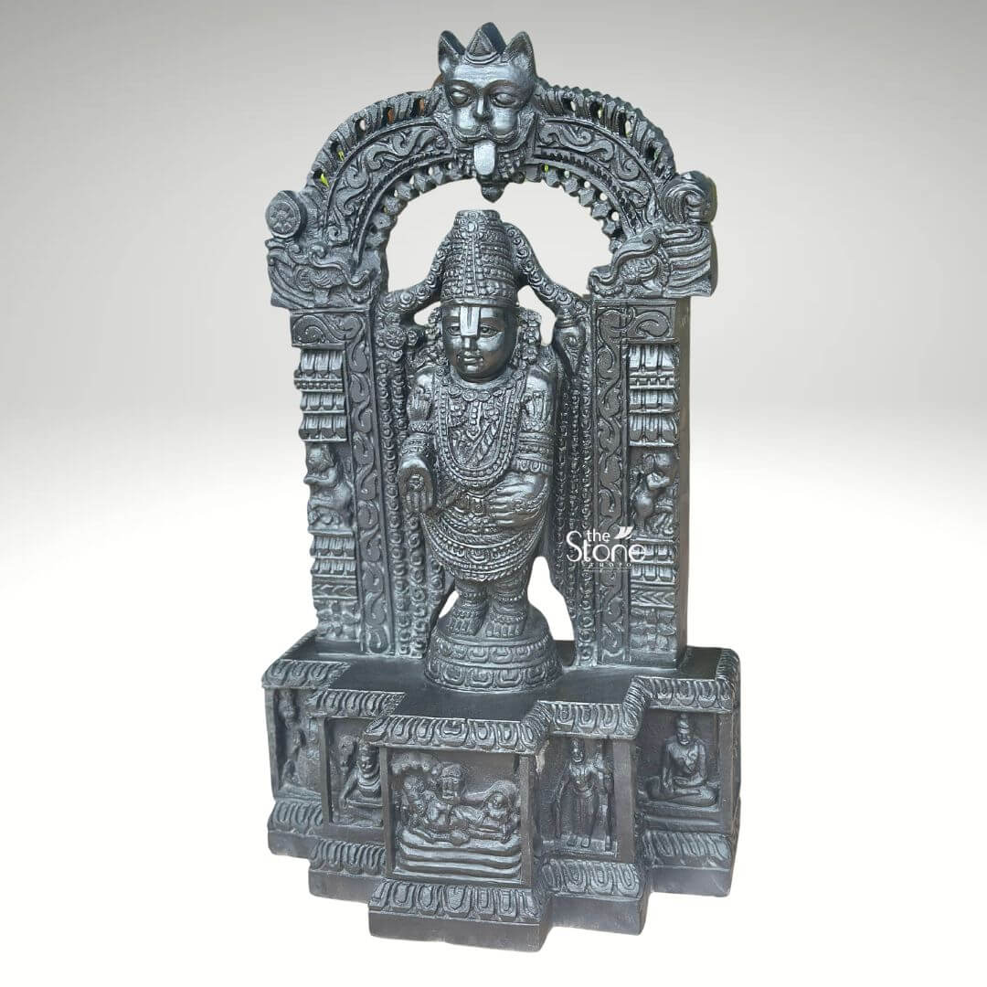 Sri Srinivasa Tirupati Balaji Statue 2.5ft 7 A detailed three-quarter view photograph captures a magnificent 2.5-foot-tall murti of Tirupati Balaji Sri Venkateswara meticulously hand-carved from a single block of dark dense blackstone. The statue depicts Lord Balaji standing in a samabhanga posture upon a lotus pedestal which is itself supported by an ornate multi-tiered rectangular base. The dark stone has a deep matte finish that accentuates the intricate sculptural details. Garuda the celestial eagle-mount is prominently carved at the apex of the elaborate prabhavali that frames the main deity. Below Garuda the arch is adorned with traditional floral and scrollwork motifs culminating in makara heads at the base of the pillars. Lord Balaji's divine form is richly adorned with finely carved traditional ornaments including multiple layered necklaces elaborate armbands bracelets and anklets. His forehead bears the distinct Vaishnava U-shaped tilak mark and his eyes are half-closed in a serene, benevolent expression. His upper right hand holds a discus while his upper left hand holds a conch shell. His lower right hand is raised in the Varada mudra and his lower left hand rests gracefully on his hip. The fine detail extends to his draped clothing which is textured to resemble silk fabric held by a jeweled girdle. The rectangular base beneath the main figure is a work of art in itself featuring intricate bas-relief panels on its visible front face. The central panel depicts the reclining form of Lord Ranganatha resting on the serpent Adisesha attended by consorts. This central scene is flanked by smaller detailed carvings of sages devotees and other deities within architectural niches. The entire sculpture from the ornate pedestal to the top of the arch reflects the traditional South Indian Hoysala and Vijayanagara architectural styles. This 2.5-foot blackstone murti is an exceptional piece of devotional art perfect for a home shrine or as a central exhibit capturing the spiritual essence and cultural heritage of the Venkateswara Temple. The photograph's clear focus on the statue's form and texture provides a comprehensive view for the viewer.