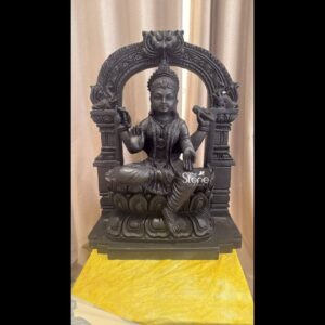 A full-body eye-level photograph of a traditional Bala Tripura Sundari idol meticulously hand-carved from a single piece of dense polished blackstone presented against a softly draped beige curtain. The statue depicts the youthful goddess seated in a graceful lalitasana pose with her right leg folded and her left leg pendant resting upon a detailed multi-tiered lotus pedestal. She is carved from a dark rich stone showcasing the natural texture and depth of the material. The goddess is adorned with intricate traditional South Indian jewelry including multiple necklaces armlets bracelets and anklets as well as a finely carved crown and elaborate ear ornaments. Bala Tripura Sundari is depicted with four arms holding symbolic attributes. Her upper right hand holds a pasha and her upper left hand holds an ankusha. Her lower right hand is raised in the abhaya mudra conveying blessing and fearlessness while her lower left hand rests upon her left knee holding a pustaka. Her face is serene and youthful with a gentle compassionate expression downcast eyes and a subtle smile reflecting deep spiritual calm and divine grace. The dark textured stone highlights the fine details of her garments ornaments and facial features. The statue is framed by a magnificently carved prabhamandala which arches over the goddess's head and incorporates traditional temple architectural elements including stylized mythical creatures floral patterns and a central kirtimukha at the apex. This ornate arch is supported by two pillars that flank the seated figure creating a sacred shrine-like enclosure. The entire composition rests on a robust rectangular black stone base which is placed on a simple yellow fabric-covered surface in the foreground. The exquisite craftsmanship and detail of the carving highlight the spiritual significance and artistic beauty of this blackstone idol of Bala Tripura Sundari making it a perfect representation of traditional temple art.