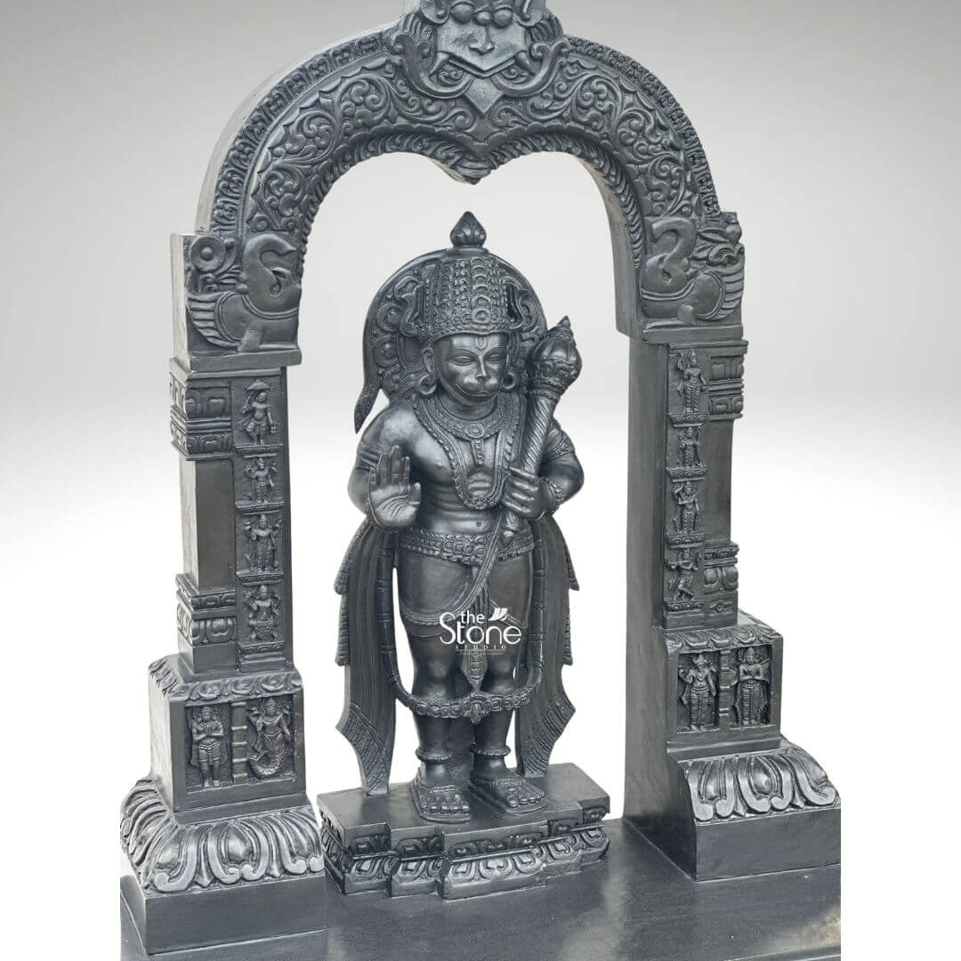 Black Stone Hanuman Statue with Arch 3ft 9 A magnificent 3-foot Traditional Hanuman Stone Idol intricately carved from deep black ston stands majestically within a grand ornate archway presented against a clean light grey background. The statue depicts Lord Hanuman in a classic standing posture radiating immense strength unwavering devotion and a vigilant readiness for service embodying the essence of a traditional deity. He is richly adorned with an elaborate crown traditional jewelry around his neck chest and arms and finely sculpted flowing garments all rendered with exceptional detail in the lustrous deep black stone. In his left hand he firmly grasps his formidable gada a potent symbol of his immense power and valor while his right hand is raised in a gesture of Abhaya Mudra offering fearlessness and divine protection to all devotees. The craftsmanship of this Blackstone Hanuman statue is truly outstanding with every minute detail meticulously brought to life adhering to traditional iconography. His facial features including his serene yet resolute expression are precisely rendered conveying his spiritual resolve and powerful persona. The subtle depiction of his musculature beneath his garments hints at his legendary strength and agility. Lord Hanuman stands on a multi-tiered black stone pedestal which is seamlessly integrated into the broader base of the entire archway structure providing both stability and enhancing its grand temple-like presence perfectly suited for a traditional idol. The statue is dramatically framed by a beautifully carved decorative arch also crafted from black stone. This arch features intricate traditional patterns elaborate floral motifs and smaller divine figures or mythological scenes meticulously carved into its supporting pillars and along the curve of the arch. These surrounding architectural elements elevate the spiritual grandeur and cultural richness of the entire piece directly reminiscent of ancient temple entrances and traditional sacred art. This impressive 3-foot Traditional Hanuman Stone Idol in Black Stone with its integrated decorative arch serves as an ideal representation for an article focusing on such a divine sculpture symbolizing unwavering loyalty courage profound spiritual strength and embodying the timeless aesthetic of a sacred traditional artifact.