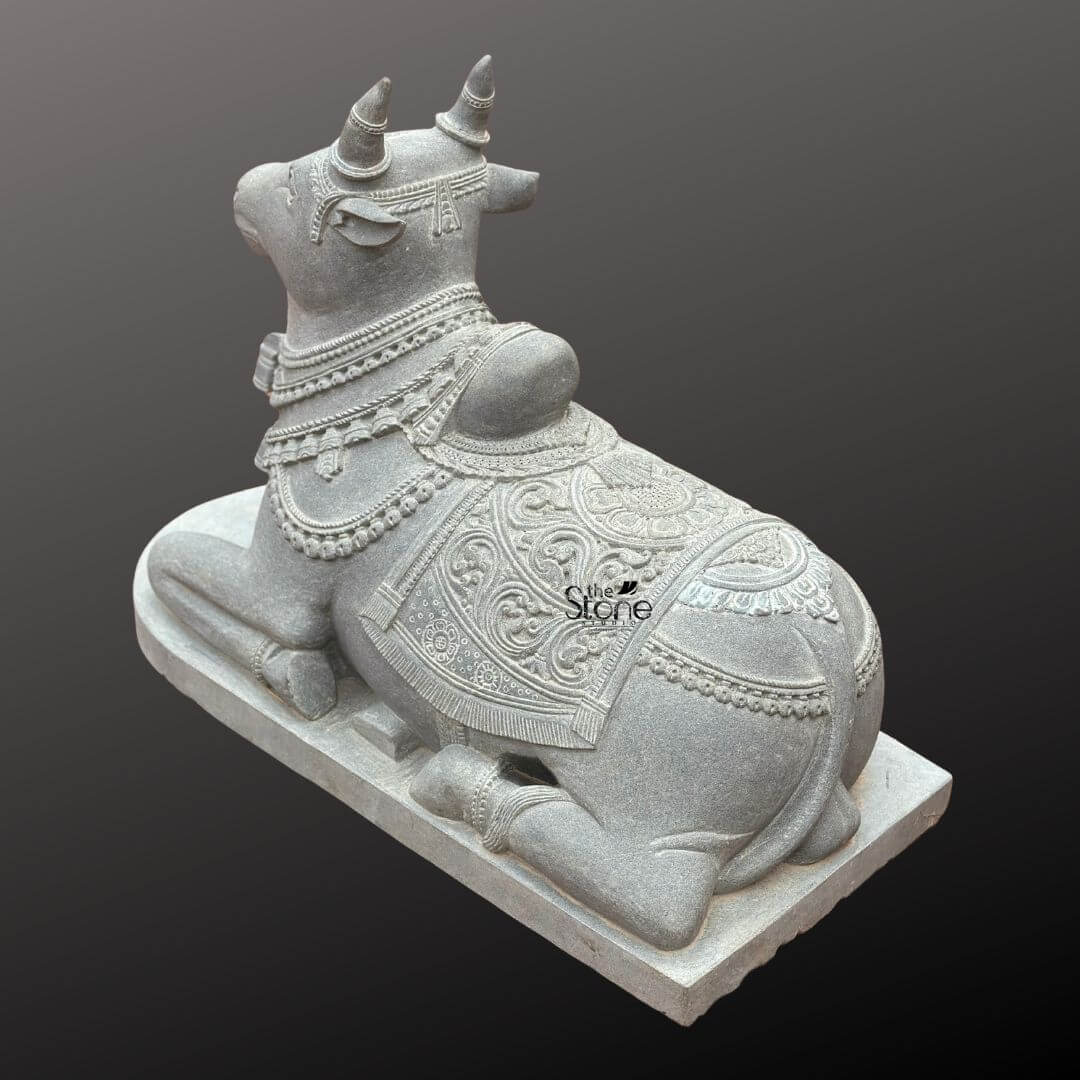 Nandikeshwara Stone Statue 45" 11 A high-angle three-quarter studio photograph showcases a magnificent hand-carved blackstone Nandi bull statue presented in a traditional recumbent pose. The sacred idol is positioned on a plain elevated rectangular stone plinth set against a smooth dark grey gradient background that emphasizes its form. The statue exhibits the dark rich and matte finish characteristic of premium blackstone meticulously worked to create both smooth surfaces and deep-relief textures. Nandi is depicted resting on its folded legs with its head turned gently to the viewer's left conveying a sense of peaceful devotion. The entire body of the Nandi statue is richly adorned with elaborate deeply carved ornamentation that highlights the artisan's skill. Its head features detailed eyes ears and stylized horns encircled by a delicate headpiece. Multiple layers of finely carved beaded necklaces and chains drape around its neck with the texture of the stone mimicking heavy temple jewelry. The sculpture conveys a sense of deep spiritual reverence through its intricate craftsmanship befitting its role as a central temple deity. The body of the Nandi is covered by a beautifully patterned saddle blanket that drapes over its back and sides. The blanket is adorned with complex deep-relief floral motifs geometric patterns and a textured border finished with detailed fringes along the lower edge. Below the blanket an intricately carved girth strap secures it ensuring the sculpture embodies the rich ceremonial decoration required for temple iconography. The overall impression is one of ancient tradition and masterful artistry making it a fitting subject for articles discussing traditional stone Nandi murtis.