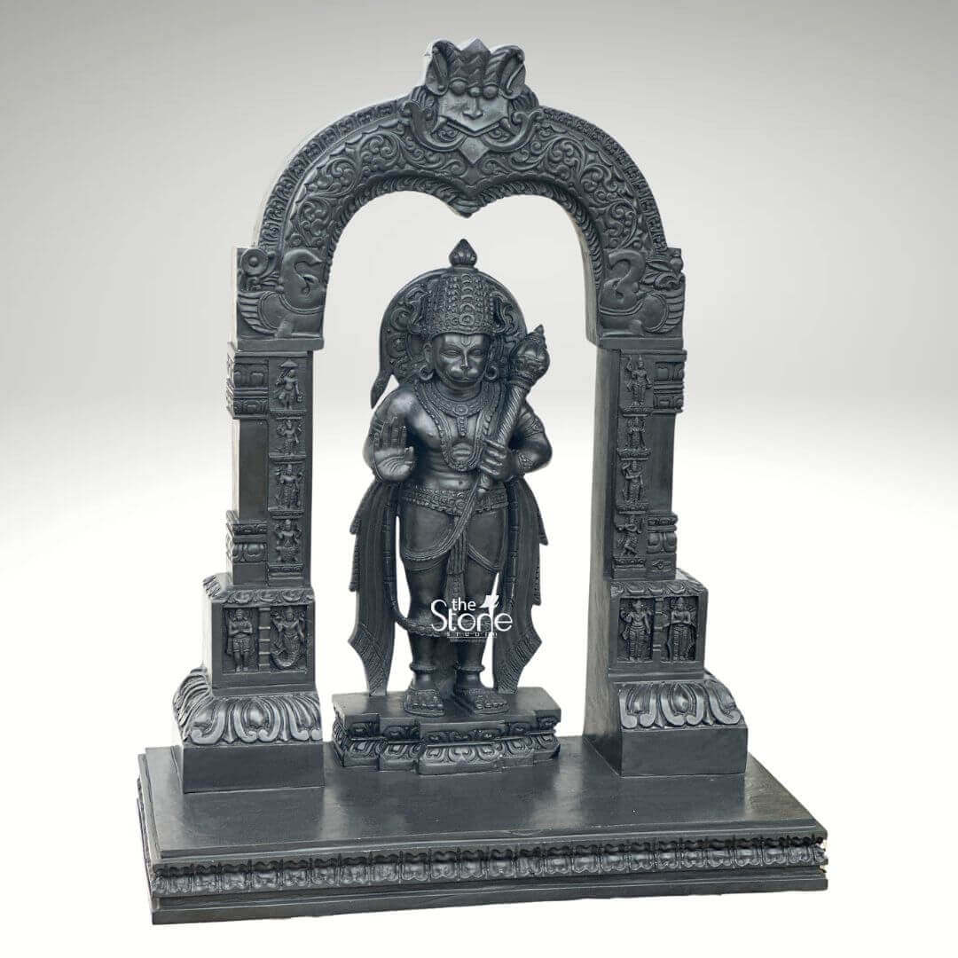 Black Stone Hanuman Statue with Arch 3ft 10 A magnificent 3-foot Veer Hanuman Black Stone Statue intricately carved stands powerfully within a grand ornate archway presented against a clean light grey background highlighting its majestic presence. The statue depicts Lord Hanuman often referred to as Veer Hanuman for his valor in a traditional standing posture radiating immense strength unwavering devotion and a vigilant readiness for service. He is richly adorned with an elaborate crown traditional jewelry around his neck chest and arms and finely sculpted flowing garments all rendered with exceptional detail in the lustrous deep black stone. In his left hand he firmly grasps his formidable gada a potent symbol of his immense power and valor while his right hand is raised in a gesture of Abhaya Mudra offering fearlessness and divine protection. The craftsmanship of this Blackstone Hanuman statue is truly outstanding with every minute detail meticulously brought to life emphasizing his heroic qualities. His facial feature including his serene yet resolute expression are precisely rendered conveying his spiritual resolve and powerful brave persona. The subtle depiction of his musculature beneath his garments hints at his legendary strength and agility perfectly suiting the epithet Veer. Lord Hanuman stands on a multi-tiered black stone pedestal which is seamlessly integrated into the broader base of the entire archway structure providing both stability and enhancing its grand temple-like presence. The statue is dramatically framed by a beautifully carved decorative arch also crafted from black stone. This arch features intricate traditional patterns elaborate floral motifs and smaller divine figures or mythological scenes meticulously carved into its supporting pillars and along the curve of the arch. These surrounding architectural elements elevate the spiritual grandeur and cultural richness of the entire piece reminiscent of ancient temple entrances and traditional sacred art. This impressive 3-foot Veer Hanuman Black Stone Statue with its integrated decorative arch serves as an ideal representation for an article focusing on such a divine sculpture symbolizing unwavering loyalty, courage profound spiritual strength and embodying the heroic essence of Lord Hanuman.