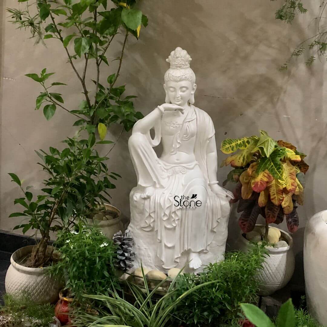 Seated Kuan Yin Statue 3ft 3 A white 3 feet fibre Guanyin statue is displayed as the central feature of a lush garden corner surrounded by layered green plants and decorative pots. The sculpture depicts Kuan Yin also known as Guanyin seated gracefully on a textured rock-style base in a calm and contemplative pose. One leg is raised with the arm resting gently across the knee while the hand is placed near the chin in a thoughtful gesture the other arm rests beside the body and one foot extends downward. The serene face softly lowered eyes delicate smile elongated earlobes and ornate crown create a peaceful expression associated with compassion mercy and spiritual tranquillity. The white Guanyin statue features flowing robes carved with long elegant folds that drape across the body and cascade over the pedestal. Fine decorative details appear around the crown neckline wrists and garment edges while the smooth white fibre finish contrasts beautifully with the natural greenery surrounding it. Tall leafy shrubs rise on the left a colourful croton plant with yellow green and reddish leaves stands on the right and smaller ornamental plants fill the foreground. White textured planters natural stones and a neutral concrete wall complete the garden setting creating a secluded and meditative atmosphere. This image illustrates how a White Guanyin Statue for Garden can become a soothing focal point in a courtyard terrace garden landscaped corner meditation area or peaceful outdoor retreat. The 3 feet fibre Kuan Yin statue blends spiritual symbolism with decorative garden styling bringing visual balance serenity and a sense of compassion to the surrounding space. Its graceful seated posture clean white finish and placement among abundant plants demonstrate how a large Guanyin garden statue can enhance both devotional environments and contemporary outdoor décor.