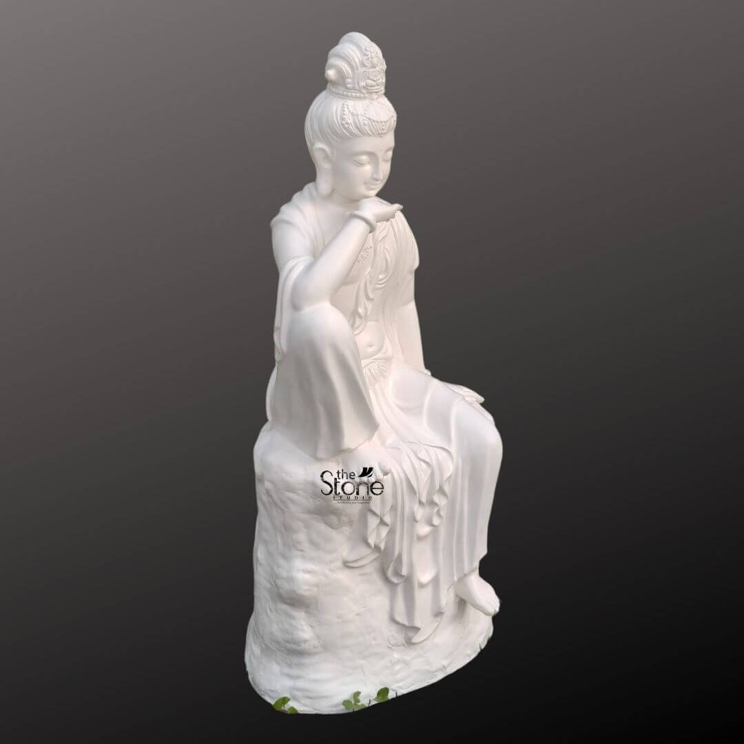 Seated Kuan Yin Statue 3ft 6 A white 3 feet fibre Kuan Yin Goddess statue is shown from a three-quarter side angle against a smooth dark grey studio background. The sculpture depicts Kuan Yin seated gracefully on a tall textured rock-like base in a relaxed and contemplative posture. One knee is raised allowing the arm to rest gently across it while the hand is positioned near the chin in a thoughtful gesture. The other hand rests softly beside the body and one foot extends downward along the front of the pedestal. Her softly lowered eyes delicate smile elongated earlobes and peaceful facial expression convey compassion serenity and spiritual calm. The White Kuan Yin Goddess Statue features an elaborately carved crown or headdress above a neatly arranged high bun along with subtle ornamental details around the forehead neckline and clothing. Long robes drape elegantly over the shoulders and body forming layered folds that cascade across the seated figure and down the textured base. The side-facing view reveals the depth of the sculpture including the raised knee curved posture flowing sleeves carefully modelled hands and graceful contours of the garment. The clean white fibre finish enhances the fine detailing and gives the statue a refined luminous appearance. This image supports an article about a White Kuan Yin Goddess Statue by clearly presenting the design material size and sculptural craftsmanship of the 3 feet fibre Kuan Yin statue. Its serene expression and meditative seated pose make it a meaningful decorative focal point for living rooms prayer areas meditation spaces wellness centres spiritual interiors covered patios or tranquil garden settings. Representing mercy compassion mindfulness and inner peace this seated Guanyin sculpture combines devotional symbolism with elegant artistic styling making it suitable for both sacred spaces and sophisticated home décor.