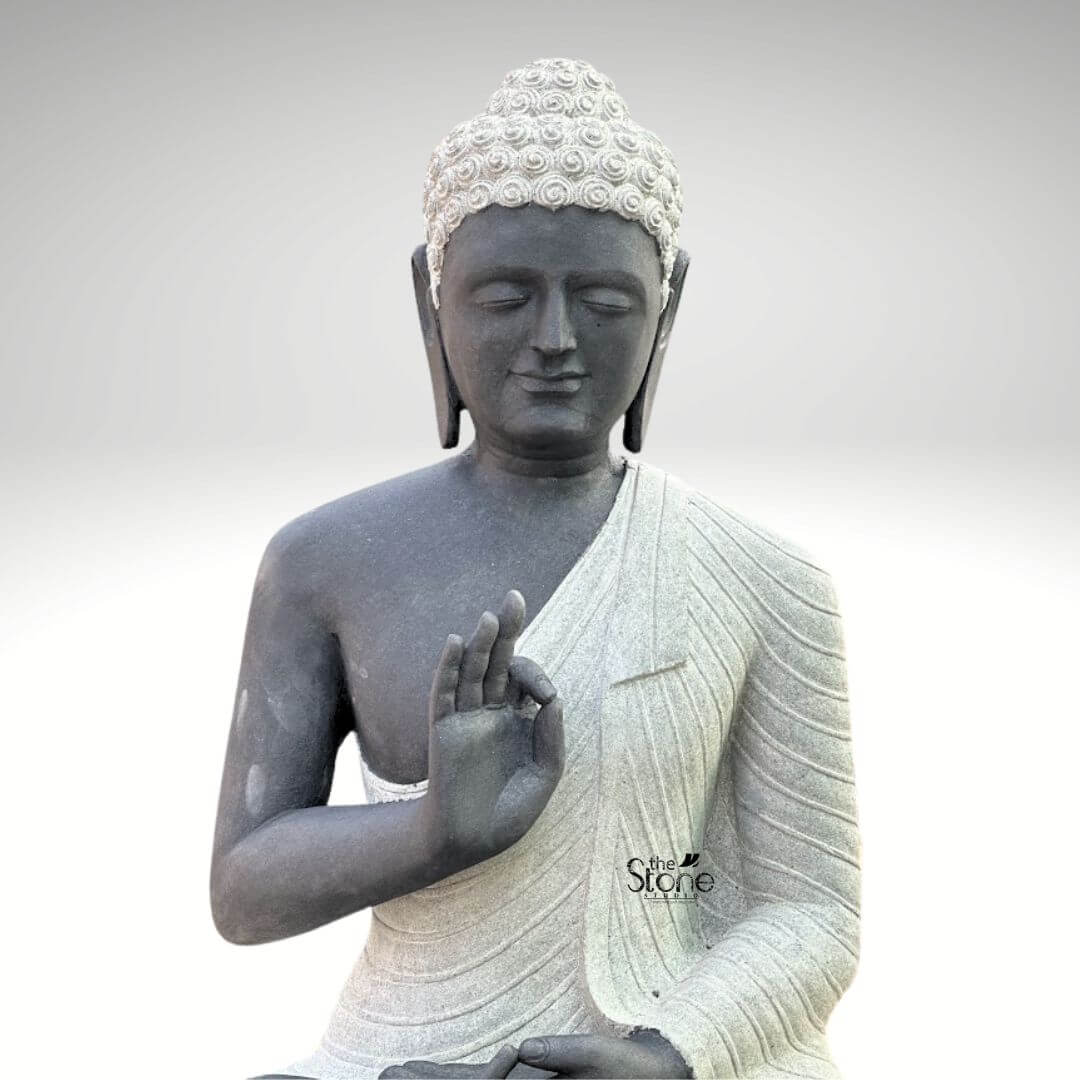 Two Tone Buddha Stone Statue for Home 3ft 6 A serene hand-carved Blackstone Buddha Stone Idol sits in a traditional meditative pose embodying the essence of Zen. The three-foot-tall statue showcases a striking two-tone finish with the Buddha's body and head carved from a deep dark blackstone exhibiting a smooth polished texture that highlights the rich dark tones. This contrasts beautifully with his monastic robes which are rendered in a contrasting lighter grey or off-white stone featuring a subtle granular texture. This distinct separation emphasizes the form of the robes as they drape over his crossed legs and shoulders adding depth and visual interest to the sacred art. The Buddha is depicted seated with his legs crossed in a full lotus position on a simple integrated dark blackstone base. His hands are gracefully positioned in the vitarka mudra symbolizing discussion and the transmission of teachings with the right hand raised and the thumb and index finger forming a circle while the left hand rests in his lap. His facial expression is calm and contemplative with downcast eyes a gentle smile and tightly curled hair leading to a cranial ushnisha all rendered with precise craftsmanship that honors traditional iconography. The overall composition evokes a sense of deep tranquility and spiritual harmony. This three-foot Blackstone Buddha Stone Idol is an ideal illustration for an article discussing a Zen Buddha Stone Idol perfectly capturing the quietude and spiritual depth associated with Zen philosophy. Its substantial size ensures it functions as a commanding focal point whether placed in a home interior as a spiritual anchor or in a garden setting. The traditional design brings a sense of sacred history and calm to any space while the durable blackstone construction ensures it is both a beautiful and a meaningful addition to its surroundings. The detailed carving and harmonious integration of elements highlight its value as a piece of sacred art.