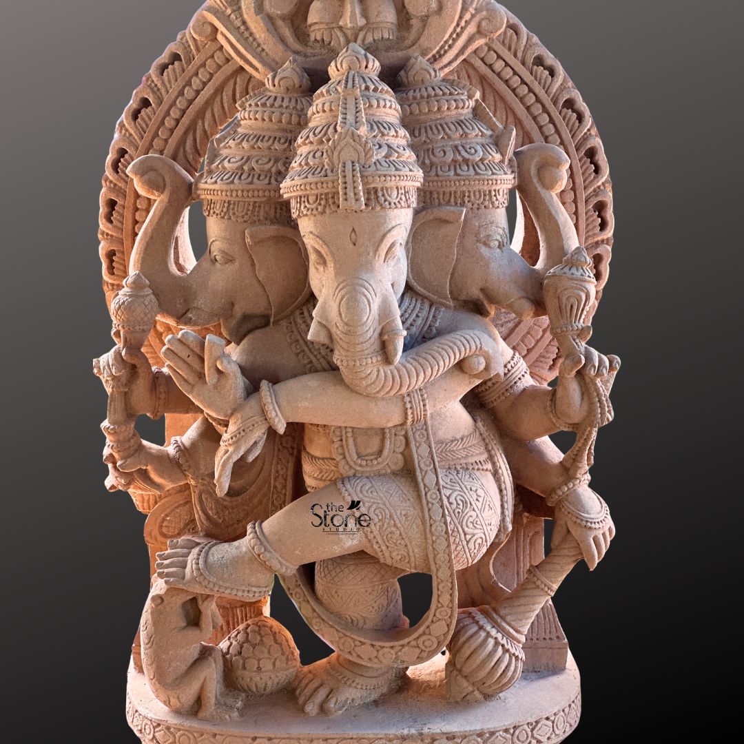 A full-view studio photograph captures a magnificent 4-foot-tall 3 Face Dancing Ganesha Stone Idol hand-carved from a single block of warm earthy sandstone. The statue is depicted in a dynamic dancing pose with Ganesha's left leg raised and bent gracefully across his body conveying a sense of divine rhythm and movement. He is adorned with elaborate traditional jewelry including multiple necklaces armlet and a sacred thread all intricately sculpted from the same durable stone. The natural porous texture and warm reddish-brown color of the sandstone are beautifully highlighted giving the sculpture a rich tactile quality. This unique murti features three heads with Ganesha's primary elephant face looking forward flanked by two identical elephant faces facing left and right all wearing ornate crowns. Ganesha is shown with eight arms each holding a symbolic attribute such as an ankusha a pasha and a modak with his trunk elegantly curved to taste a sweet from his lower left hand. The sculpture rests on a finely detailed, multi-layered lotus throne, grounding the monumental figure. Behind him a large ornate halo with traditional patterns frames his head adding to the divine aura. At Ganesha's feet a small figure of his vahana the mouse is depicted looking upwards in reverence. This image serves as an exemplary representation of traditional Indian stonemasonry and is a perfect illustration for an article discussing the significance of a 3 Face Dancing Ganesha Stone Idol. The photograph highlights the exceptional detail and the imposing presence of the 4-foot sandstone statue making it easy for a devotee or collector to appreciate its spiritual energy and artistic grace. The clean neutral grey background ensures that the viewer's focus remains entirely on the sculpture's form texture and complex iconography conveying the significant role such a murti plays in a home shrine temple or garden setting.