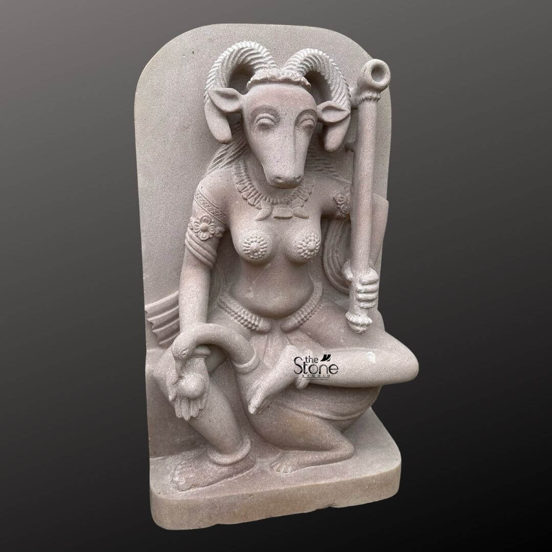 This is a centered full-view photograph of a meticulously hand-carved off-white sandstone statue of the Yogini Vrishanana standing two feet tall and presented against a seamless dark grey studio background that fades to black. The statue is carved in a high relief panel style depicting the Yogini a powerful female deity in a complex seated posture with her right leg bent and her foot resting on her mount a recumbent bull. She is sculpted with a human female body and the distinctive head of a horned goat creating the unique Vrishanana form. Her expression is calm and focused gazing forward and she is adorned with deeply carved traditional jewelry including multiple necklaces armlets and a detailed waistband all rendered directly from the stone. The Yogini Vrishanana statue is depicted with four arms showcasing complex iconography. Her primary right hand holds a small fruit or offering while her primary left hand rests gracefully on her raised left knee. In her upper right hand she holds a water pot and in her upper left hand, she firmly grips a ritual staff topped with a symbolic disc. The statue's off-white sandstone material has a finely textured matte finish that emphasizes the high level of detail and the deep reliefs of the carving conveying a sense of age and reverence appropriate for this type of ancient-style art. The statue stands on a simple integrated sandstone base. The composition captures the complete form of the Yogini and her mount presenting the specific iconographic details that define the Vrishanana form. The soft even directional studio lighting illuminates the sculpture from the front and slightly to the side casting gentle shadows that define its three-dimensional form and highlight the rich tactile quality of the sandstone surface. The image accurately represents the two-foot scale of the piece making it a suitable representation for an article discussing this specific ancient Yogini form.